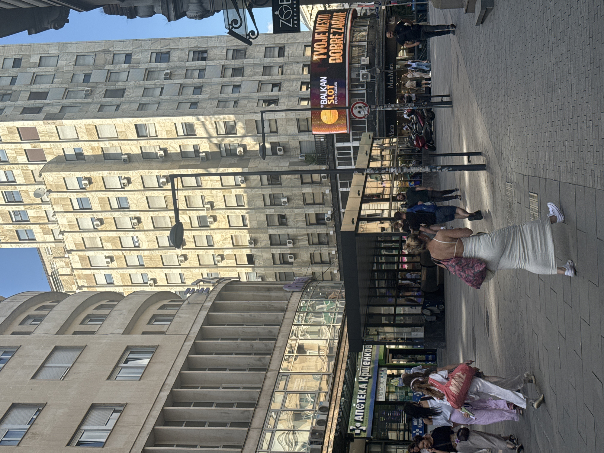 The image captures a street scene in Belgrade, Serbia, specifically in front of the Albanija Palace. The architecture features two prominent buildings: a curved, Brutalist-style building on the left with repetitive vertical elements and rows of windows, and a taller, rectangular building with a stone facade on the right, also adorned with numerous windows and air conditioning units.
In the foreground, a group of people are walking on a paved sidewalk. A woman in a long, white, form-fitting dress and white sneakers is walking away from the camera, carrying a tote bag with a floral pattern. To her left, another woman in a white tank top and a long, striped skirt walks with a large red tote bag. Other individuals, dressed casually, are also navigating the pedestrian area.
A large digital billboard is mounted on the side of the building on the right, displaying advertisements, including one for "BALKAN SLOT" and another with text in what appears to be Serbian. Below the billboard, the facade of a MaxMara store is visible.
The environment is urban, with shops and businesses lining the street. The lighting suggests it is daytime, with shadows indicating the sun's position. The overall impression is a bustling urban street in a commercial district of Belgrade.