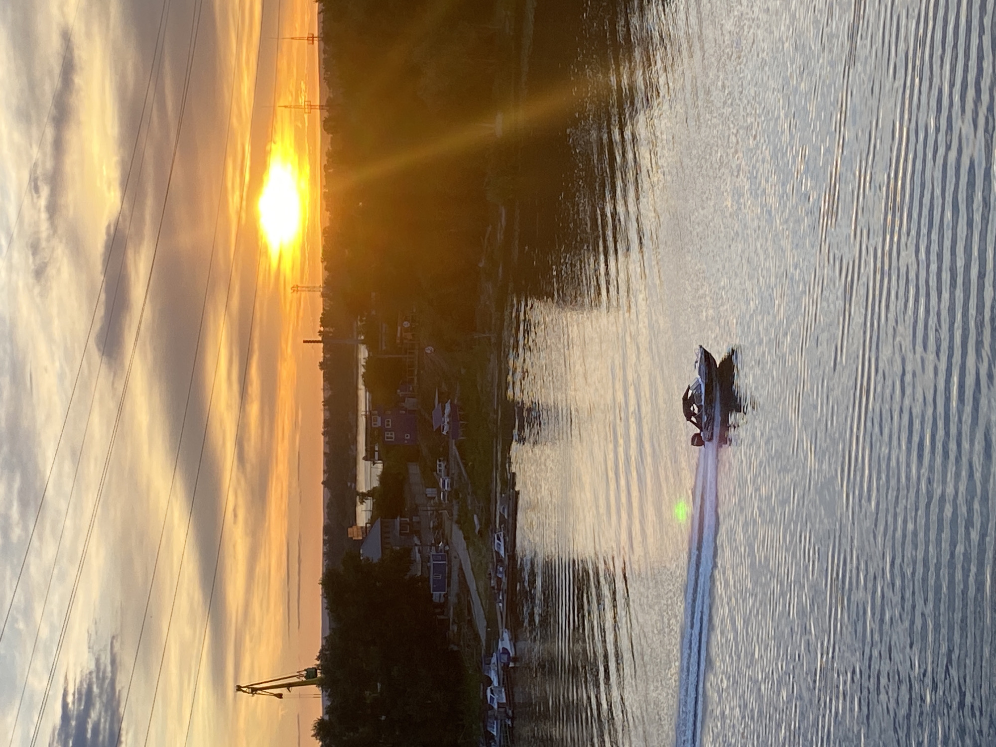 The image captures a serene sunset over the Volga River near Novokuybyshevsk, Russia. The sky is ablaze with hues of orange and yellow, with the sun prominently positioned near the horizon. Power lines crisscross the sky, and a large crane stands as a testament to industrial activity in the area.
On the riverbank, several boats are docked. A single speed boat is the focal point of the water, creating a white wake as it moves across the surface. The water reflects the warm tones of the sunset, creating a tranquil and picturesque scene. The lush green foliage lining the riverbank adds a touch of natural beauty to the industrial landscape.
