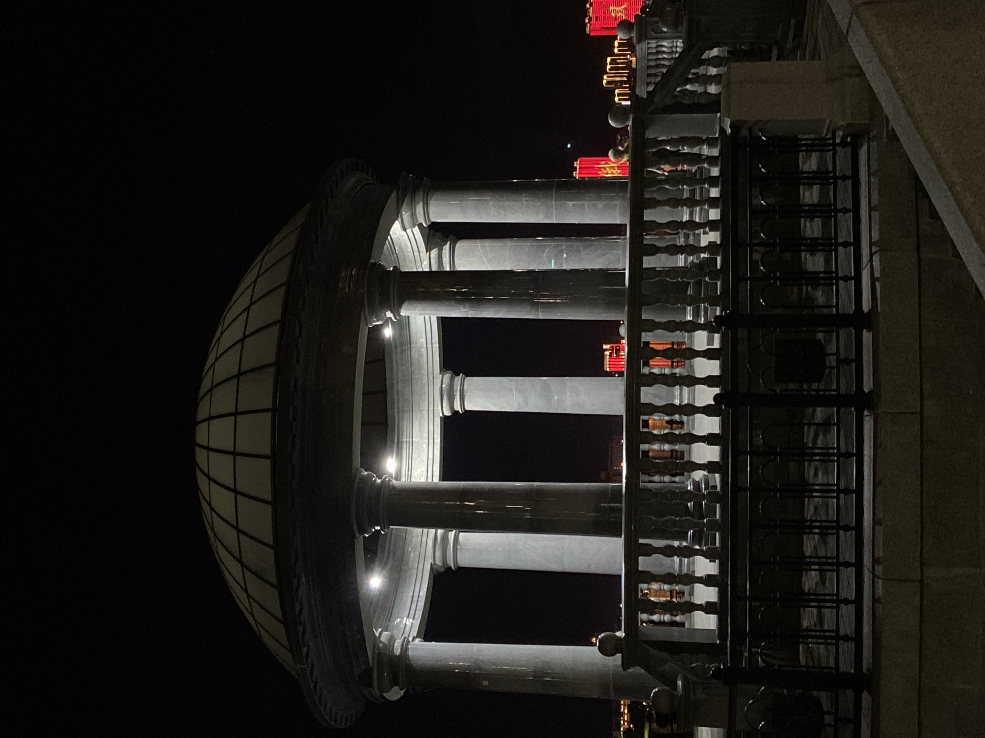 The image shows a nighttime view of a domed pavilion with classical columns, likely in Blagoveshchensk, Russia. The pavilion is illuminated from within, casting light on its marble columns and the surrounding paved area. A decorative railing, made of stone balusters and a wooden handrail, encloses the pavilion. Beyond the railing, a dark fence is visible. In the background, illuminated buildings with red lighting are visible, suggesting an urban setting. The sky is dark, indicating it is nighttime. There are no people clearly visible within the pavilion itself, but a few figures appear to be in the distance. The overall scene is one of architectural grandeur set against a nighttime urban backdrop.