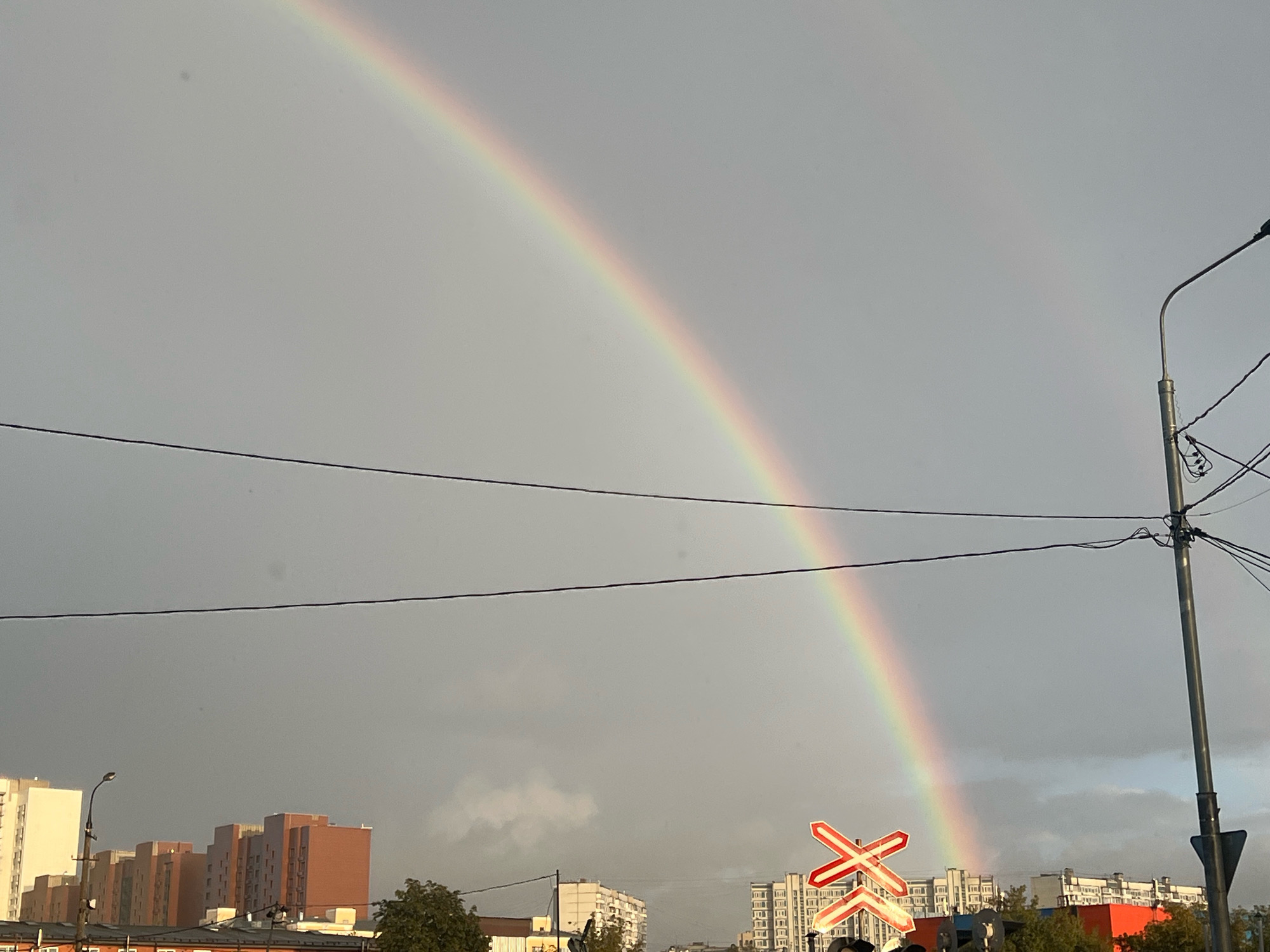 A rainbow arches across a cloudy sky, with a faint secondary rainbow visible above. Below the rainbow, residential buildings with various colored facades are visible, interspersed with trees. A railway crossing sign, featuring red and white diagonal stripes, stands to the right of the frame. Electrical wires crisscross the sky, and a lamppost with a dark fixture is also present. The scene appears to be an urban environment, possibly in Moscow, Russia, given the architectural style of the buildings and the presence of a railway crossing.