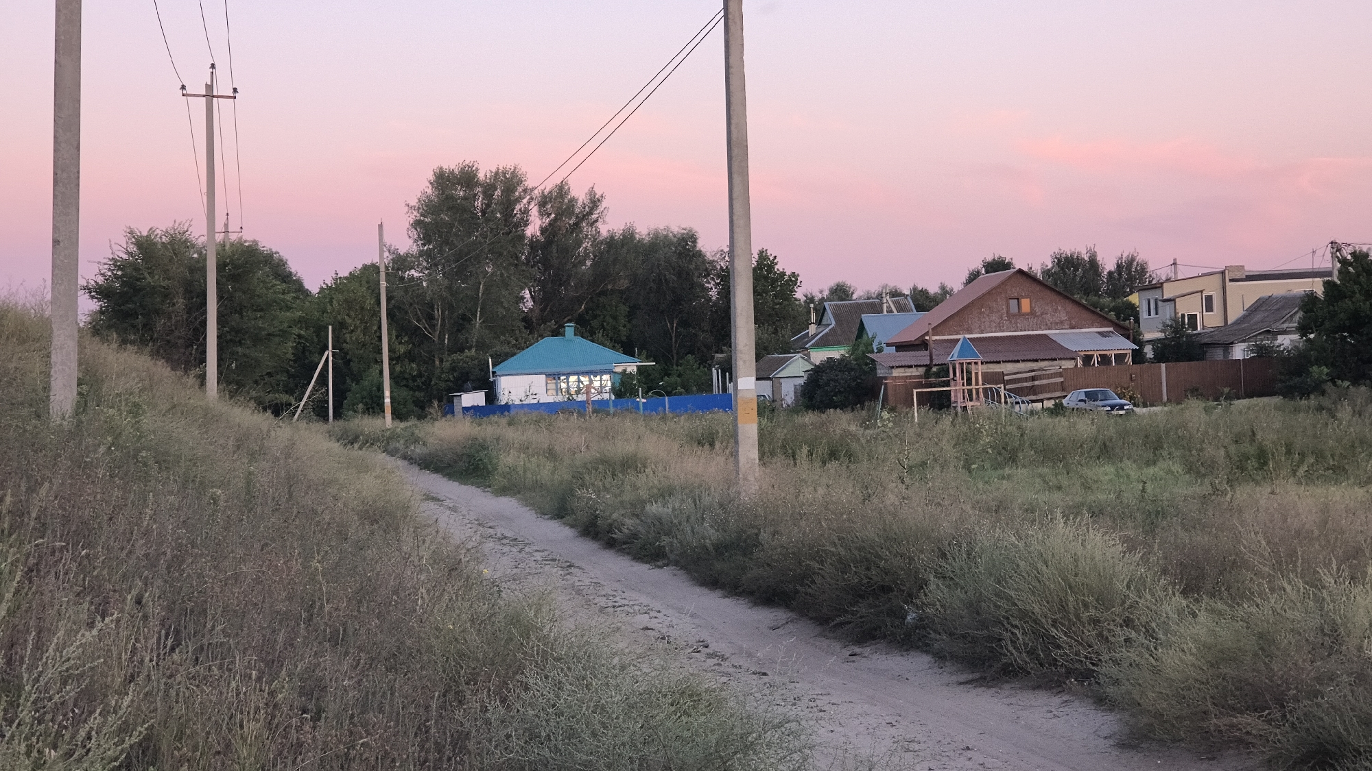 The image depicts a rural scene in Mikhaylovka, Russia, during twilight. A dirt road leads through tall, dry grass and weeds, curving towards a cluster of houses in the background. The sky is a soft gradient of pink and pale orange, characteristic of sunset. Several utility poles with wires are visible along the road and in the distance.
The houses are of various sizes and architectural styles, suggesting a residential area. One prominent house features a bright turquoise roof and is surrounded by a blue fence. Another larger house has a brown roof and an attached children's play structure. A dark-colored car is parked in front of one of the houses. Trees and foliage surround the houses, adding to the natural, somewhat wild, aesthetic of the setting. The overall impression is one of a quiet, late evening in a small Russian town.