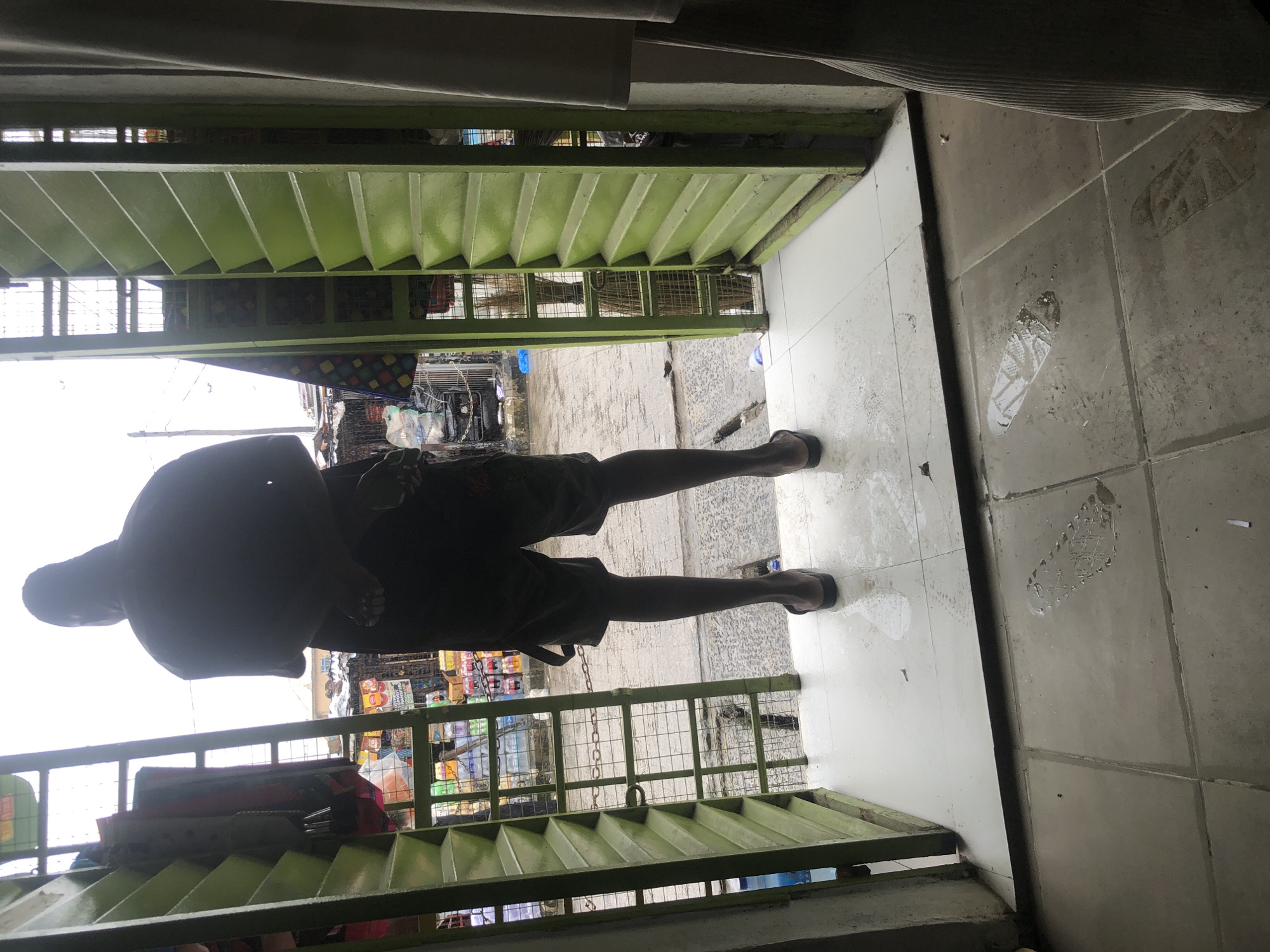 The image depicts an outdoor scene in a busy market area, likely in Ado, Nigeria. The perspective is from inside a storefront, looking out through an open doorway framed by green louvered shutters. A person, viewed from the back, stands with their hands clasped behind their back, wearing shorts and flip-flops. They are silhouetted against the bright, overcast sky.
Beyond the person, the market unfolds. Stalls with various goods, including bottled water and other products, are visible. A chain-link fence separates the immediate area from the market stalls. The ground outside appears wet and there are visible footprints and water puddles on the tiled floor of the storefront, suggesting recent rain. A broom can be seen leaning against a rack behind the fence. The overall impression is one of a bustling, possibly humid, urban market environment.