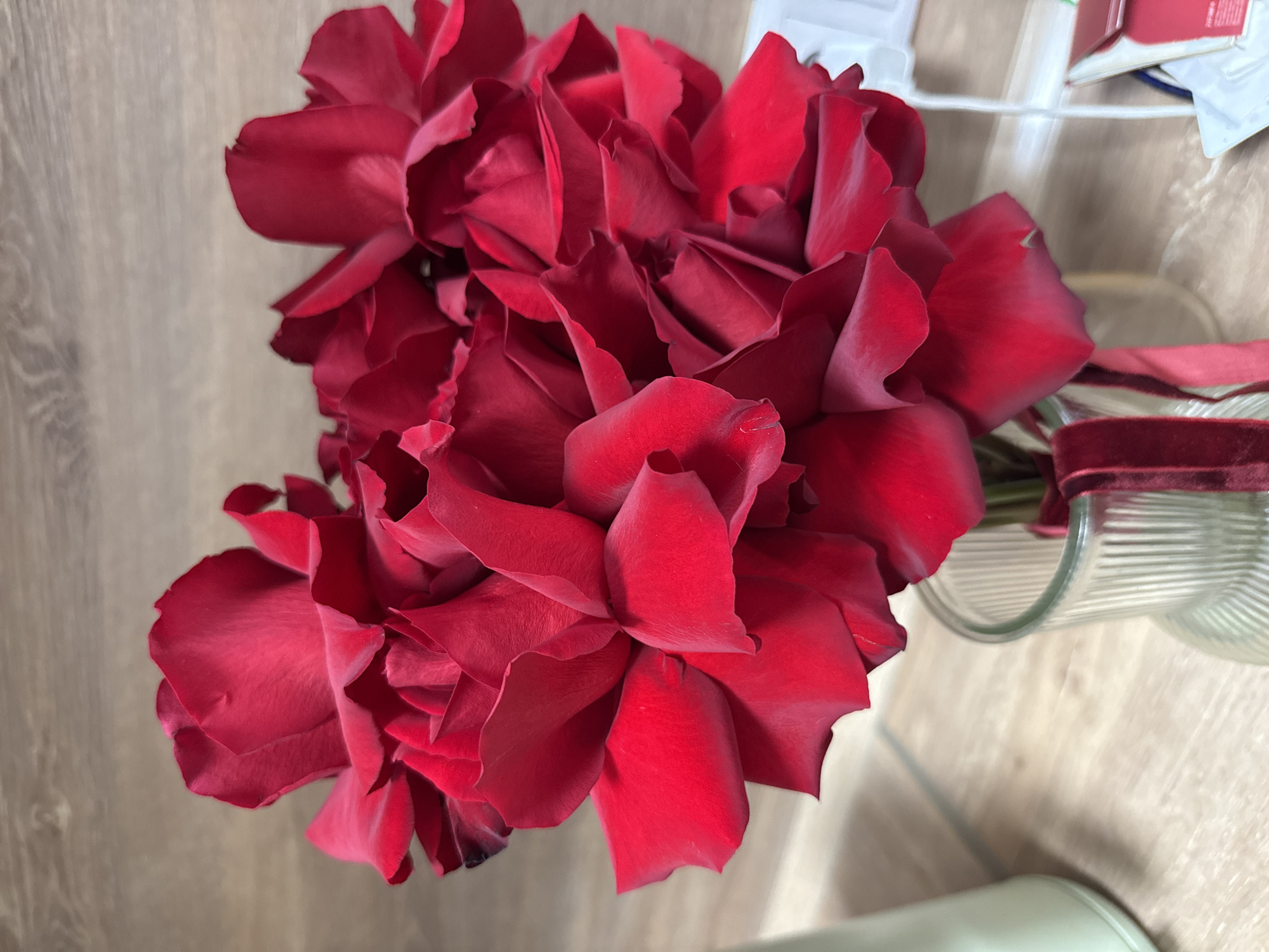 An elevated, close-up photograph displays a dense bouquet of deep red roses. The numerous roses dominate the frame, showcasing their rich, velvety petals, many of which appear fresh with natural curls and gentle creases. Some petals exhibit very minor dark spots along their edges.
The roses are held in a clear glass vase, visible on the right side of the image. The vase features a distinctive ribbed texture and has a dark red, possibly velvet, ribbon tied around its neck.
The bouquet is placed on a light-colored wooden surface, which could be a table or floor, revealing a subtle wood grain pattern. The background, particularly in the upper right, contains blurry household items, including papers and parts of boxes or containers, suggesting an indoor setting. A light green, out-of-focus object is partially visible in the lower right corner. The lighting is soft and appears natural, illuminating the roses without casting harsh shadows. No activity is taking place, and there is no visible text. The scene is located in Izhevsk, Russia.