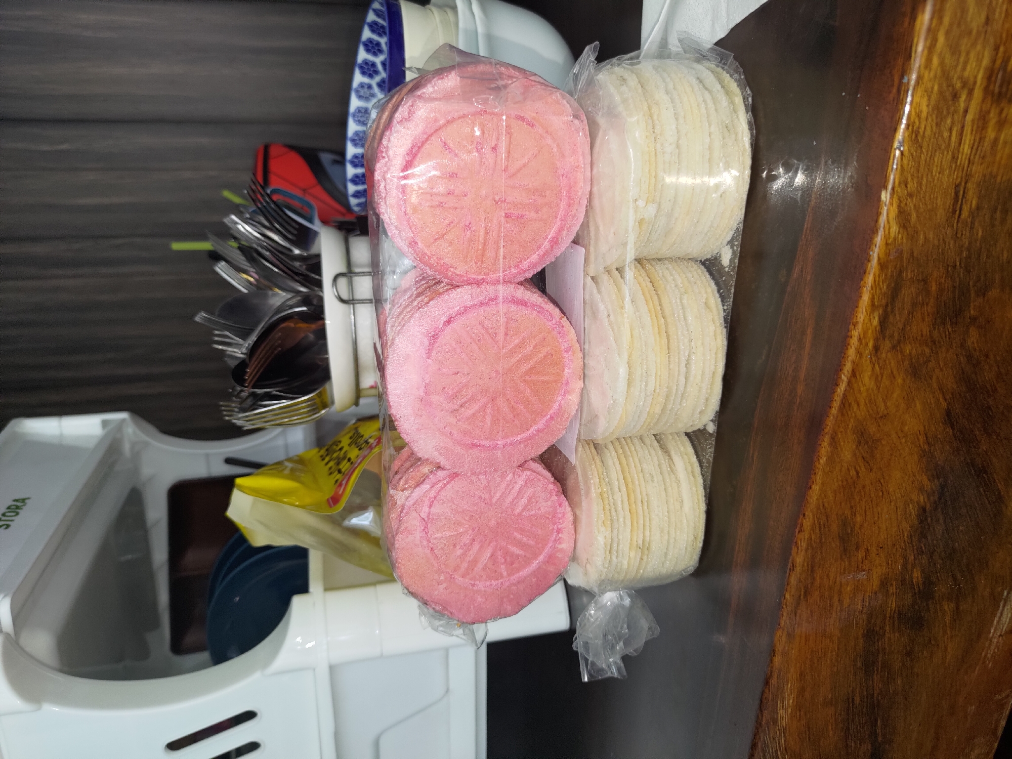 The image shows a countertop in Depok, Indonesia, featuring a white dish drying rack on the left, holding blue plates and a yellow package. A collection of silverware is visible in a white container behind the dish rack. In the foreground, three packages of round, flat food items are stacked on a dark wooden surface. The top two packages contain pink, circular items with embossed patterns, possibly cookies or crackers. The bottom package contains stacked, light-colored, circular items, likely tortillas or similar flatbreads. The lighting suggests an indoor setting, possibly a kitchen or dining area.