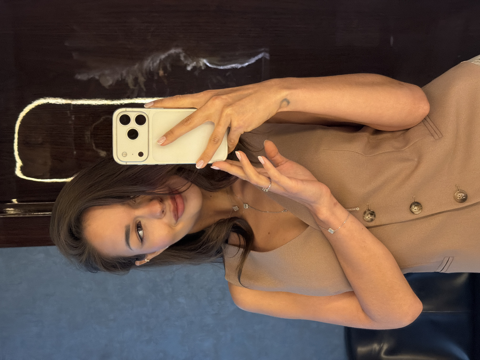 A young woman is captured in what appears to be a mirror selfie, holding a light-colored smartphone with a visible triple camera setup in her right hand. The image is oriented vertically with her head towards the bottom left.
The woman has dark hair and a soft, slight smile on her face, looking towards the camera. She is wearing a sleeveless, light brown or tan top with large, round, dark buttons down the front. Her attire suggests a casual yet put-together style.
Notable details include several pieces of jewelry: a delicate silver necklace with small, decorative charms (one resembling a clover, others square-shaped), a slender silver bracelet with a small, rectangular charm on her left wrist, and rings on her fingers. Her fingernails are manicured in a French tip style. A small, dark tattoo, possibly a crescent moon, is visible on her left forearm.
The background consists of two distinct areas. Behind her head and shoulders, there is a dark, highly reflective surface, possibly polished wood or a dark mirror, showing some light reflections. Below her, the surface appears to be a textured, grayish-blue wall or panel. A black, possibly leather, surface is visible beneath her right arm. The setting appears to be indoors. No visible text is present in the image. The location context provided is Moscow, Russia.