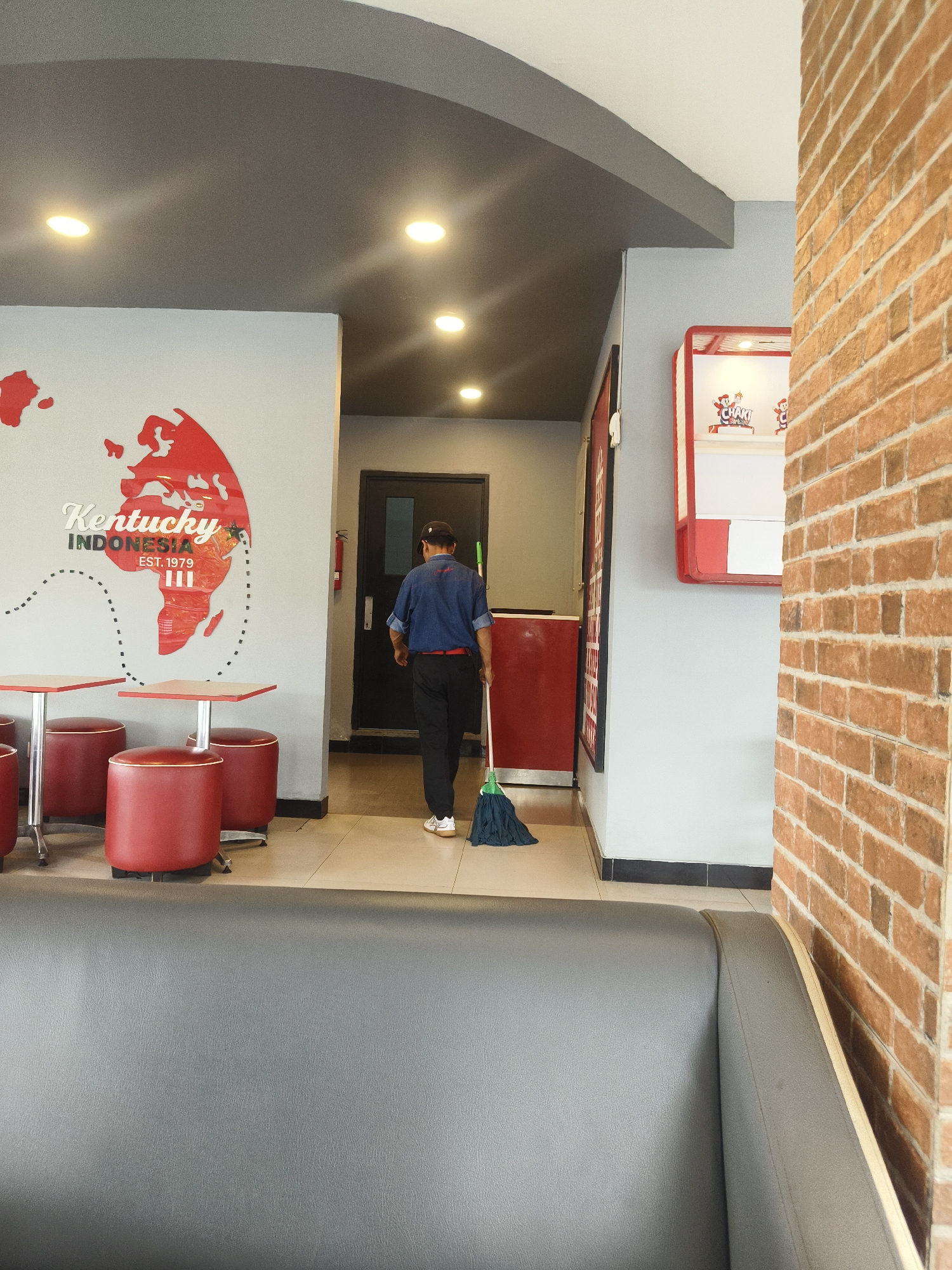The image depicts the interior of a fast-food restaurant, likely a Kentucky Fried Chicken branch in Denpasar, Indonesia. The setting features a grey ceiling with recessed lighting, and a white wall adorned with a red map graphic and text indicating "Kentucky INDONESIA EST. 1979". To the left, there are red circular stools and small square tables. A man in a blue denim shirt, dark pants, and a baseball cap is seen walking away from the camera towards a doorway, holding a green-handled mop. The floor is tiled with a light-colored pattern. To the right, a brick wall is visible, and further to the right is a red display case with branding that appears to be related to the restaurant. A dark grey sofa occupies the foreground, suggesting a seating area for customers. The overall impression is of a clean and functional commercial space.