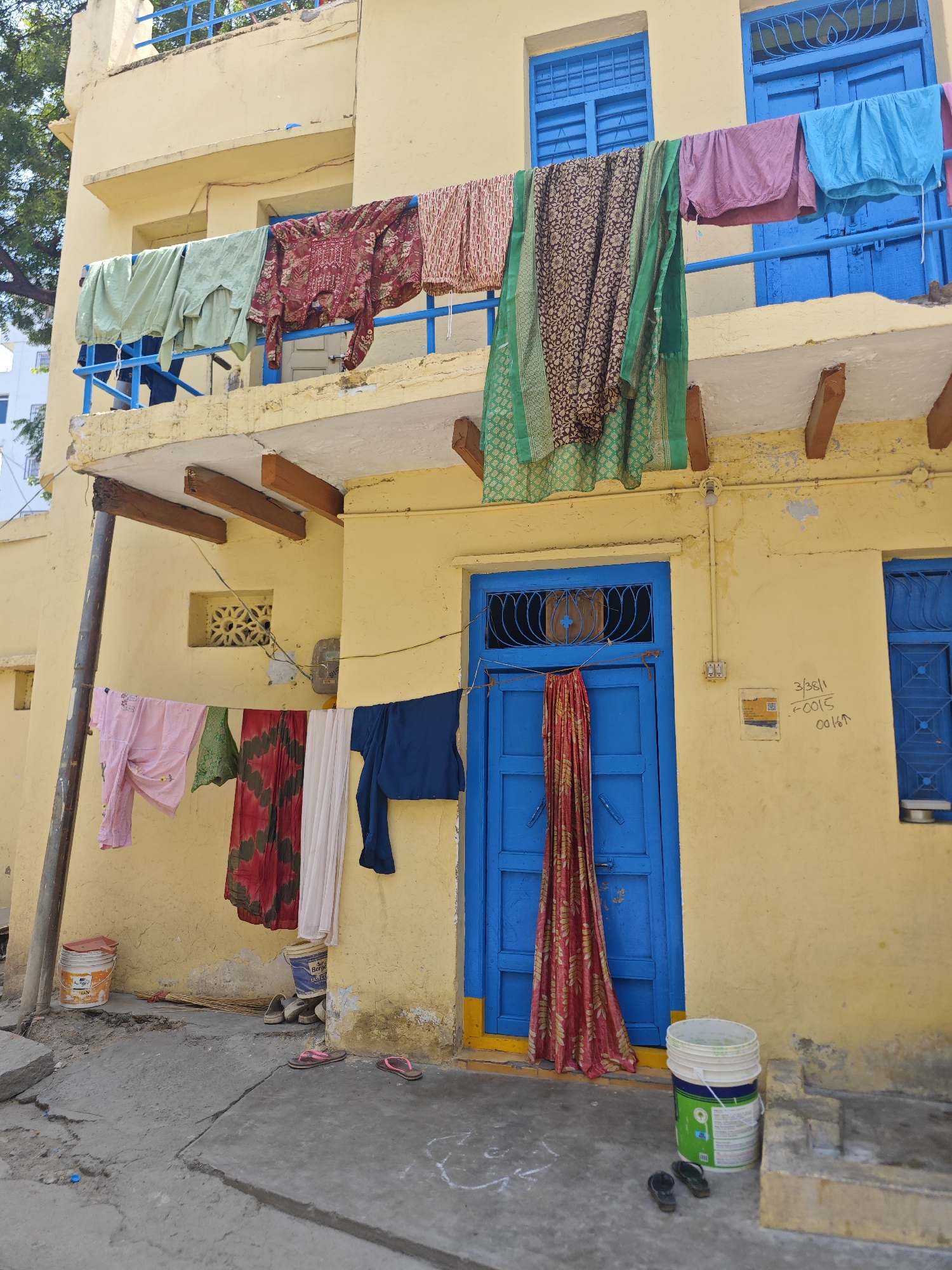 The image depicts the exterior of a two-story building in Pedda Kammavaripalli, India, with laundry hung to dry. The building's facade is painted yellow, with blue-painted window frames and doors. The first floor features a balcony with a blue railing where various garments, including a green garment, a patterned maroon shirt, and a floral-patterned fabric, are suspended on a clothesline. Below the balcony, another clothesline displays pink, green, red, white, and blue clothing items. A vibrant red and gold patterned fabric is also draped across the blue front door. On the ground level, a broom, two pairs of flip-flops, and two buckets are visible. One bucket is orange and white with "asian" branding, and the other is white with green and blue branding, labeled "berger". A small, decorative vent is present on the yellow wall to the left of the door. The ground is paved with concrete, showing cracks and unevenness. The scene is captured during daylight, casting shadows and highlighting the bright colors of the clothing and building.