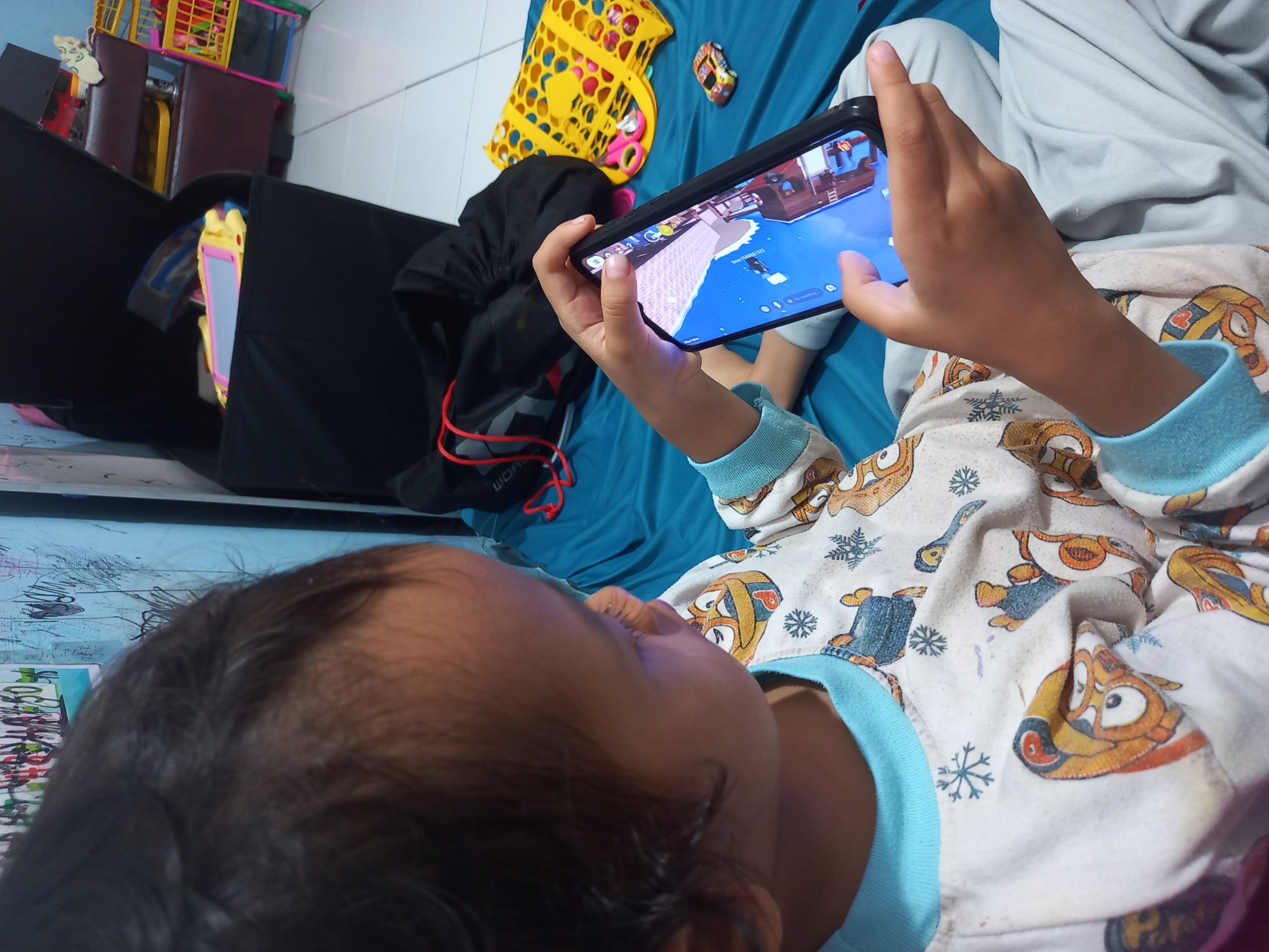 A child, seen from a close-up, over-the-shoulder perspective, is lying on a blue mat and playing a game on a smartphone. The child is wearing pajamas with a cartoon character print. The background includes a black storage bin with toys inside, a small toy car, and a yellow textured object. The environment appears to be an indoor residential setting.