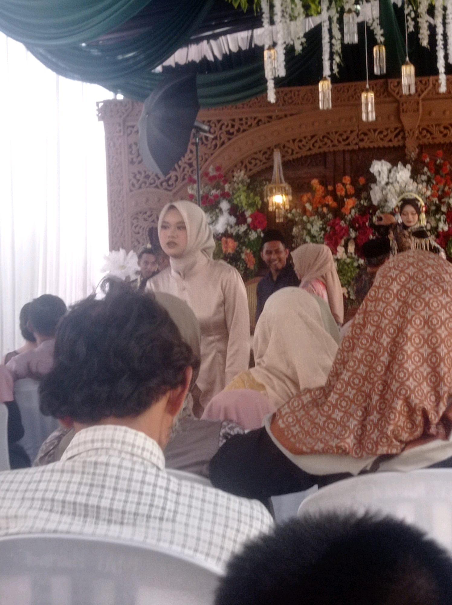 The image depicts a wedding reception ceremony, likely in the city of Ngemplak, Indonesia. In the foreground, a person wearing a plaid shirt is seated, their back to the camera. To the left, a woman in a beige hijab and dress is standing. In the midground, several individuals are visible, including a man with a dark shirt and a woman wearing a traditional gold and black wedding outfit and veil, with flowers behind her. The background features a decorated wooden archway, green draped fabric, and hanging lights.