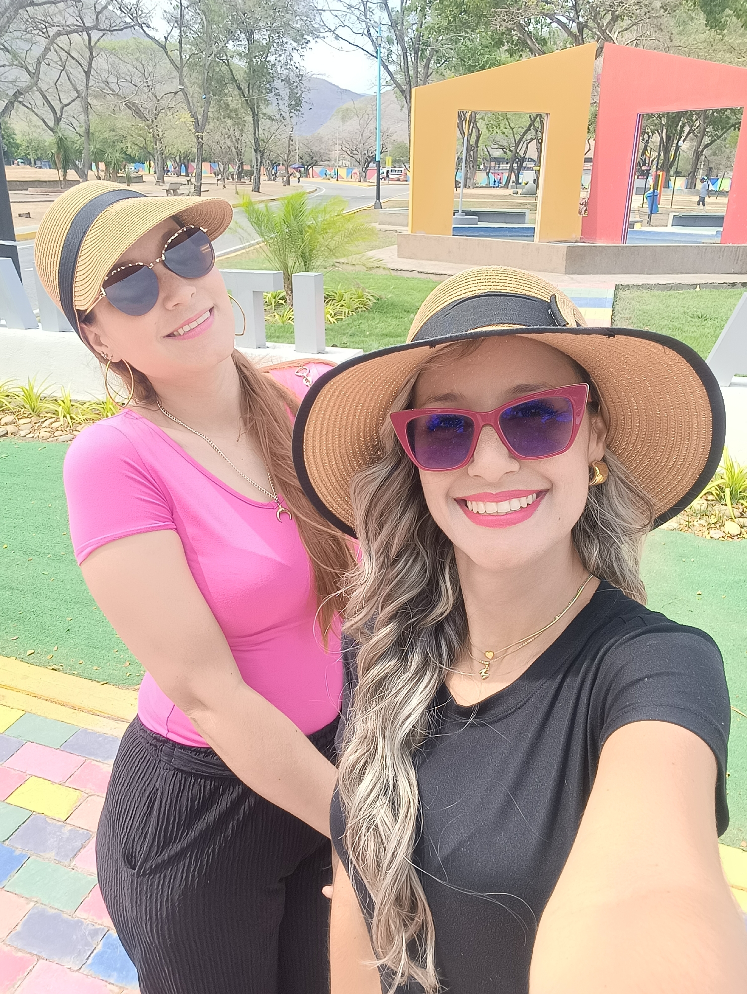 Two adult females are captured in a close-up, selfie-style photograph within an outdoor public park.
Female 1, positioned to the left and slightly behind, wears a pink short-sleeved top and black pleated pants. She wears a straw visor with a black fabric band, dark sunglasses with gold-colored frames, and gold-colored hoop earrings. A gold chain with a crescent moon pendant is visible. Her hair is light brown. She smiles, looking towards the camera.
Female 2, positioned to the right and foreground, wears a black short-sleeved t-shirt. She wears a wide-brimmed straw hat with a black fabric band, pink cat-eye sunglasses with dark lenses, and gold-colored stud earrings. Two gold-colored necklaces, one with a small heart pendant and another with a letter pendant (appearing as 'M'), are visible. Her hair is wavy and highlighted. She smiles, looking directly towards the camera, and holds the capturing device.
The immediate foreground features a multicolored tiled pathway with a checkerboard pattern of pink, yellow, blue, and white, adjacent to a flat green surface. The background identifies the location as Plaza de La Victoria in La Victoria, Venezuela. Visible elements include white block letters forming parts of the word 'LA VICTORIA', landscaped areas with small green plants, and a paved road. Distinctive architectural features are present: two large, angular, open frame structures, one yellow and one red, standing on a raised platform. A smaller blue structure is visible behind the red frame. A white van is partially visible on the road. Deciduous trees and trees with green foliage line the park, with mountains visible on the distant horizon under an overcast or hazy sky. The scene is illuminated by natural daylight.