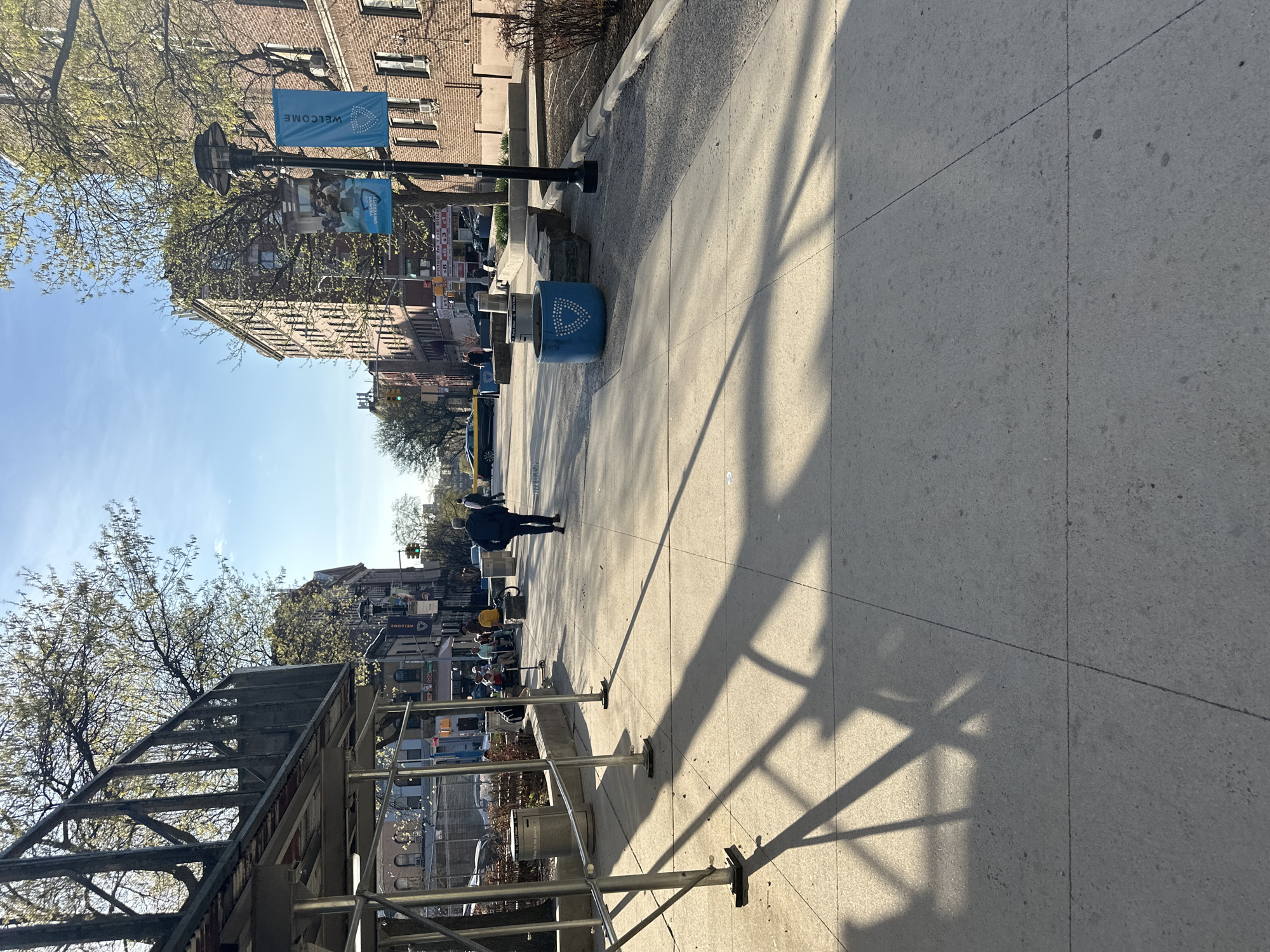 The image depicts a sidewalk scene in what appears to be New York City. The sidewalk is concrete, with visible shadows from a scaffolding structure and trees. A person walks down the sidewalk, heading toward the distance. Buildings line the street on both sides, with scaffolding visible on the left. Blue banners with the word "WELCOME" are attached to a lamppost. Trash cans and other street furniture are present. The sky is blue with scattered clouds.