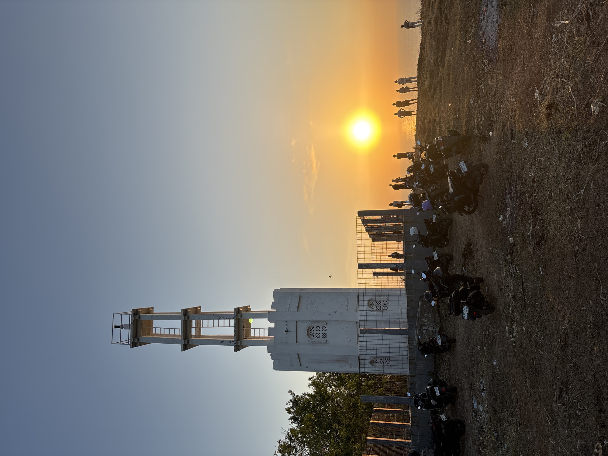 In Kuta Selatan, Indonesia, at sunset, a white tower with crenellated battlements and a ladder extending to its top stands on a hill overlooking the ocean. The tower is partially surrounded by a metal fence and adjacent to trees. A group of people are gathered on the edge of the hill, silhouetted against the bright sun setting over the water. In the foreground, a collection of parked motorcycles is visible on the uneven, dry ground. The sky is clear with a gradient of blue to orange hues.