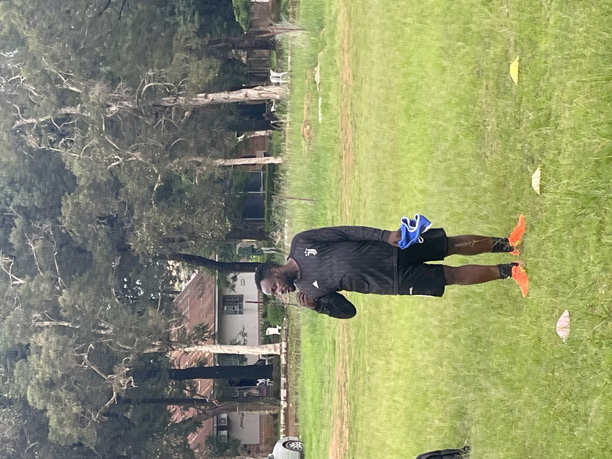 A Black man, appearing to be an athlete, stands on a grassy field, oriented vertically in the image. He is wearing a dark, long-sleeved athletic shirt with a subtle striped pattern and a white logo on the left chest, and black shorts. He holds a pair of blue and white gloves in his left hand, and his right hand is near his chest, possibly adjusting his shirt. His feet are clad in bright orange athletic shoes. The setting is an outdoor field with lush green grass, interspersed with fallen leaves, some of which are yellow and light pink. In the background, there are trees with intricate branches and a few buildings, including what looks like a residential house with light-colored walls and a red-tiled roof, and another building with brickwork. The overall impression is of a training session or a break during an outdoor sports activity. The time of day appears to be daytime, with diffused lighting, suggesting overcast weather. The location is identified as Jos, Nigeria. There is no visible text beyond the logo on the man's shirt.