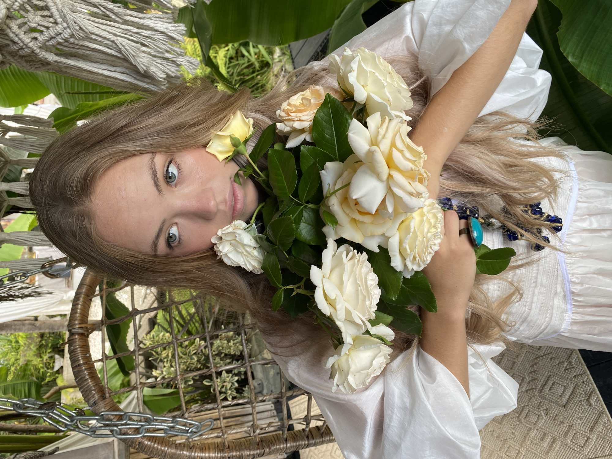 A young woman with long, blonde hair and blue eyes is lying down, holding a bouquet of cream-colored roses. She is wearing a white dress and is adorned with jewelry, including a turquoise ring and a necklace with blue beads. The setting appears to be outdoors, possibly in a garden, with lush green foliage visible in the background. She is seated in a woven chair that is suspended by chains, suggesting a relaxed and serene atmosphere. The lighting suggests it is daytime, with natural light illuminating the scene. The woman's expression is calm and contemplative. There is no visible text in the image.