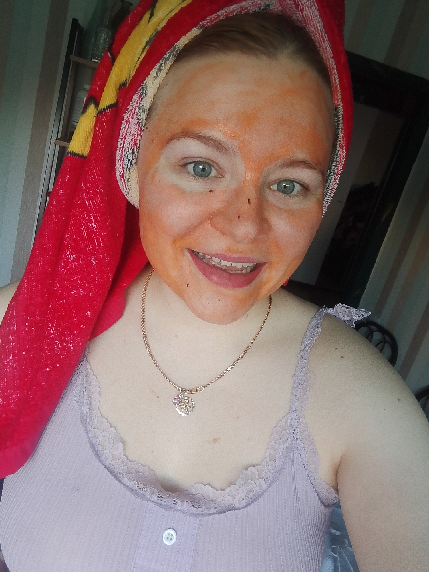 A young, light-skinned female with blue eyes is centered in the frame, appearing to take a selfie. She has an orange-colored facial mask applied to her face. A red and yellow patterned towel is wrapped around her head. She is wearing a light purple camisole with lace trim, and a gold necklace with a circular pendant. She is smiling, revealing her teeth.
The background shows a room with a striped wall. A shelf unit is visible on the left, and a partially visible black frame, possibly a mirror or picture frame, is on the right.
There is no information in the image to suggest that this photo was taken in Izhevsk, Russia.
