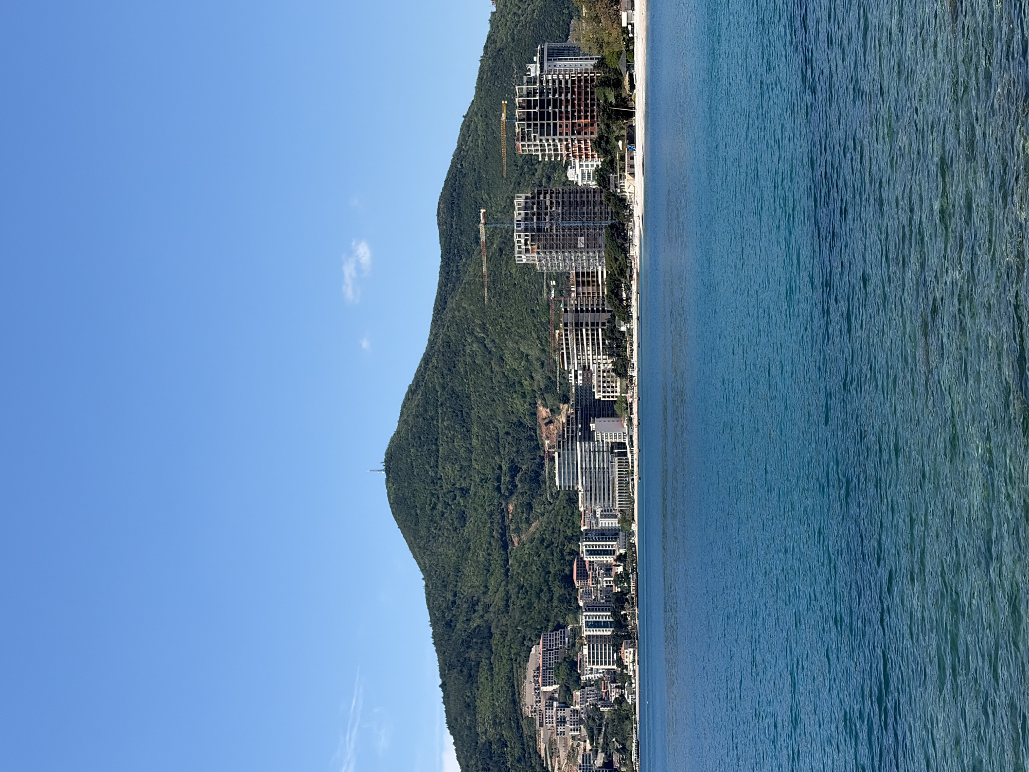 The image depicts a coastal cityscape, likely near Budva, Montenegro. The scene is oriented with the sky on the left, the shoreline in the center, and the sea on the right.
A verdant, mountainous hill dominates the landscape behind the city. Numerous buildings of varying sizes are clustered along the coastline and ascend the lower slopes of the hill. Construction cranes are visible among some of the buildings, suggesting ongoing development.
The sea appears calm with visible textures on its surface. A narrow strip of beach is situated between the buildings and the water. The sky is clear and blue.