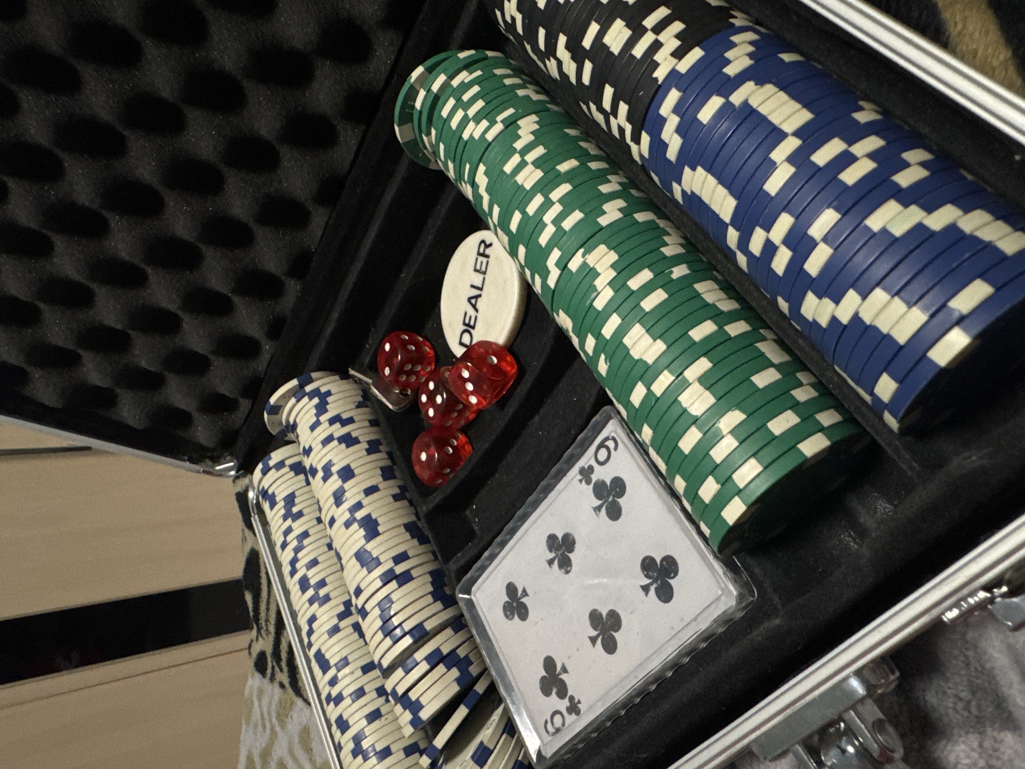 An open poker set case contains stacks of green, blue, and white poker chips, dice, a dealer marker, and a six of clubs playing card. The case has a black foam interior. The location of the image is unknown and cannot be determined from the image.