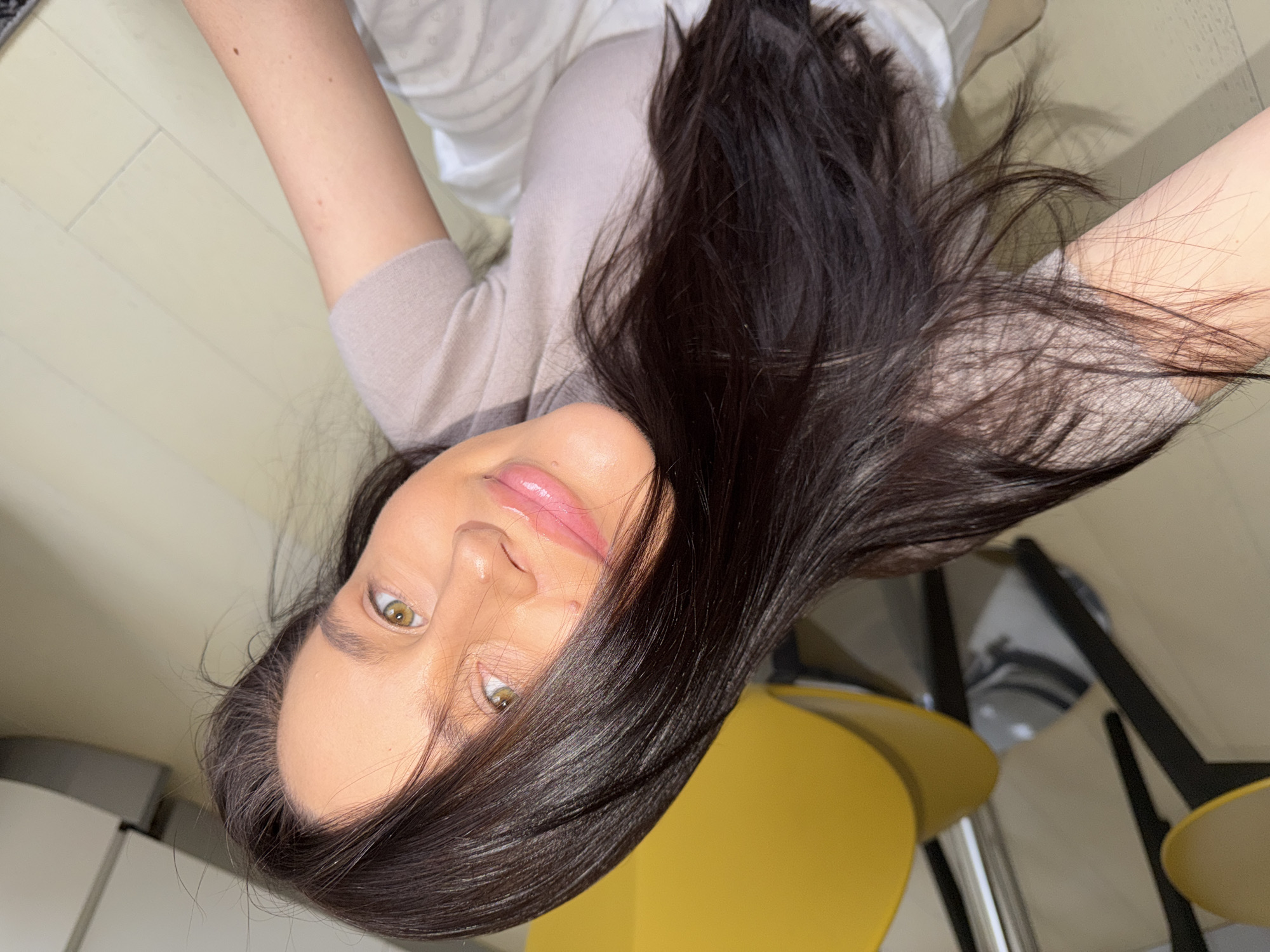 An overhead photograph, intentionally rotated, features a young woman lying down, looking directly upwards at the camera. She has long, dark brown hair, which is fanned out around her head. Her face is visible, characterized by striking green or hazel eyes, dark eyebrows, and glossy pink lips with a subtle, serene expression. Her skin appears smooth, and some moles or freckles are visible on her arm.
She is dressed in a light-colored, possibly beige or light grey, knitted top, with a white garment visible beneath it. The background suggests an indoor setting, likely a home in Khimki, Russia. A light-toned wooden or laminate floor occupies much of the lower frame. On the right side, parts of a bright yellow chair and black chair legs are visible. To the left, a light-colored surface, possibly a cabinet or counter, can be seen. The overall lighting is bright, indicative of daytime. There is no specific activity depicted beyond posing for the photograph. No visible text is present in the image.