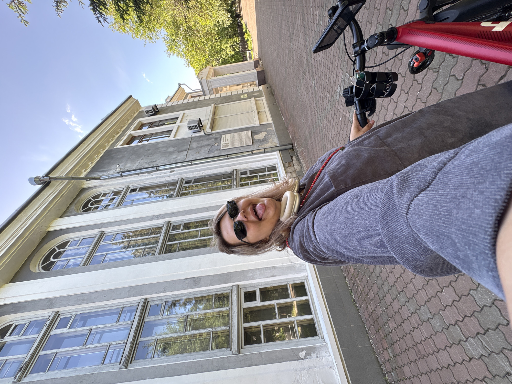 A medium shot captures a young woman on a bicycle in front of a building. She is facing the camera, sticking her tongue out and wearing sunglasses and headphones. The building behind her has tall, arched windows and is painted in shades of grey and white. The ground is paved with red and grey bricks. To the right of the woman, the handlebars of her red bicycle are visible, along with a phone mount and a cup holder. Trees and a clear blue sky are visible in the background. The overall scene suggests a sunny day in an urban environment. The building's architecture suggests it could be a public building, possibly a school or administrative center. The presence of trees and paved walkways implies a park or campus setting within Birobidzhan, Russia.
