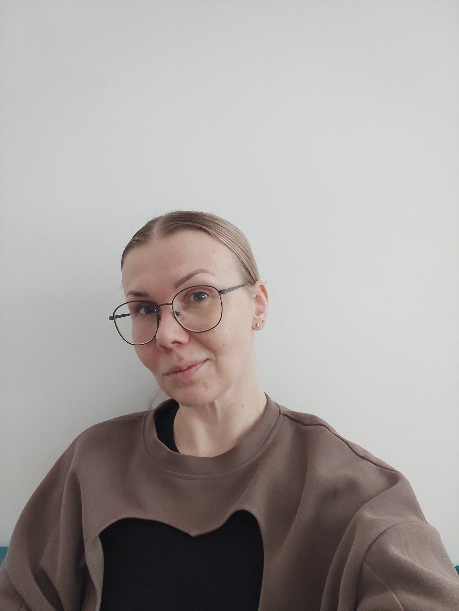 A woman with light skin and blonde hair pulled back is centered in the frame, wearing glasses with thin black frames. Her attire consists of a brown, long-sleeved sweatshirt over a black top, which is visible at the neckline and through a cutout design in the front of the sweatshirt. She is looking directly at the camera with a slight smile. The background is a plain white wall, which takes up a significant portion of the upper frame. The scene appears to be indoors, and the lighting is bright and even. The location is in the city of Unknown, Unknown.