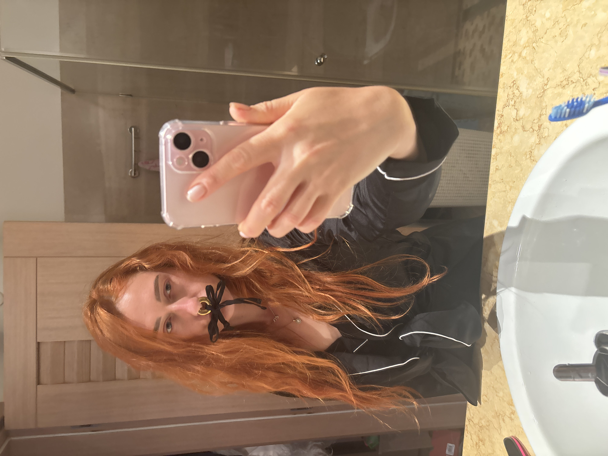 An individual with long, wavy red hair is captured in a mirror selfie within a bathroom setting in Budva, Montenegro. The person, visible from the chest up, wears a black top with white piping and a silver-colored necklace with a small pendant. A prominent gold-colored ring pierces the nasal septum, from which a black ribbon extends, tied in a bow over the mouth, functioning as a gag. The individual holds a light pink smartphone, enclosed in a clear case, in their right hand, positioned to reflect the scene. The visible environment includes a white ceramic sink and a light brown marble countertop in the foreground. In the reflection behind the individual, a light-colored wooden door is situated to the left, and a glass-paneled shower enclosure is to the right. A light brown tiled wall and a chrome towel bar with a small, light-colored towel are also discernable.