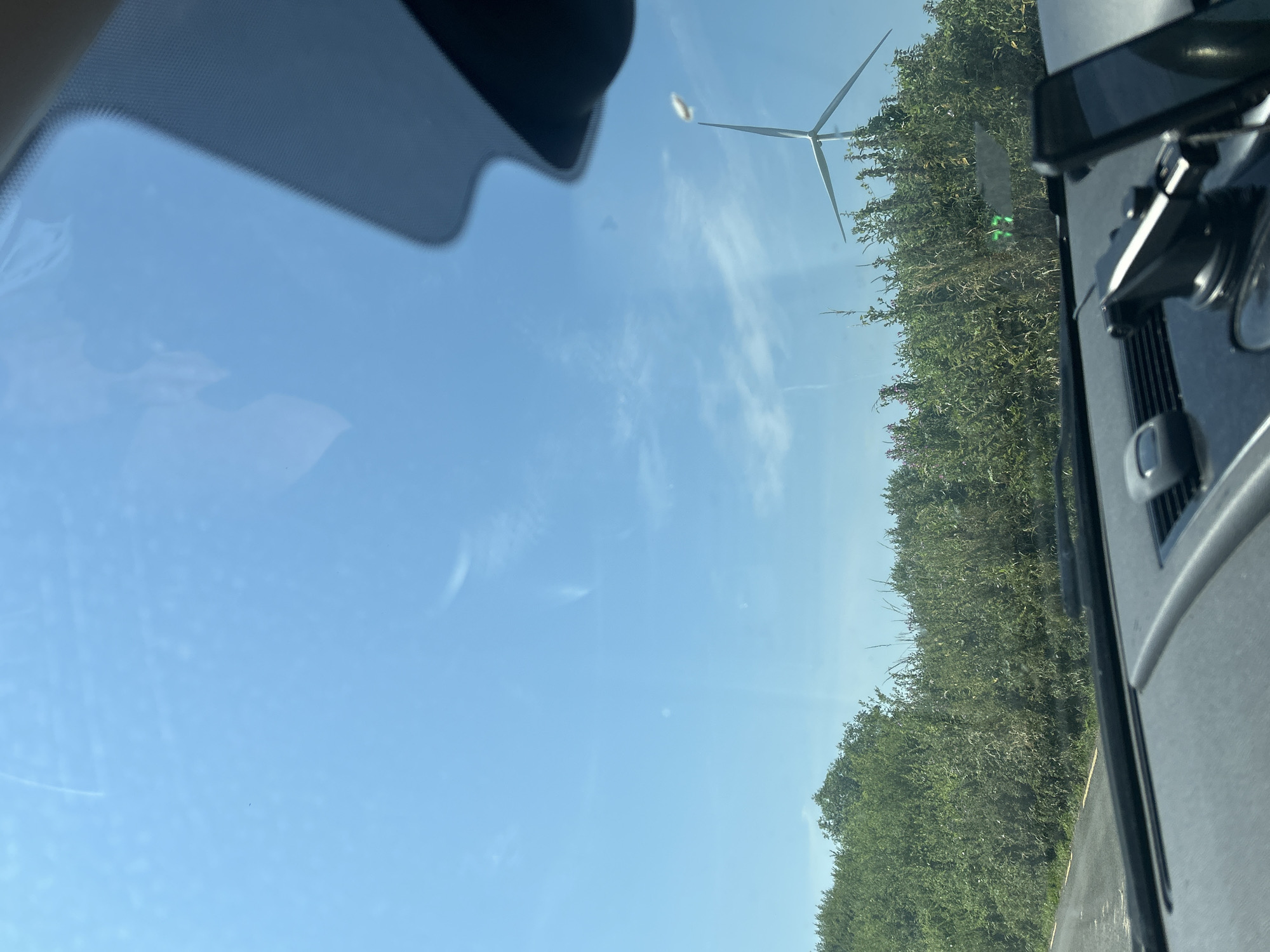 The image shows a view from inside a vehicle in Cross of The Wood, Ireland. The foreground is dominated by the dashboard of a car, including a phone mount and a phone. The windshield reflects the sky and a portion of the car's interior. Beyond the windshield, a landscape is visible under a clear blue sky with some wispy clouds. A dense line of green foliage forms a barrier across the middle ground. To the right of the foliage, a wind turbine stands tall, with its blades extended. A road with a yellow line is visible on the left side of the image, running alongside the foliage.