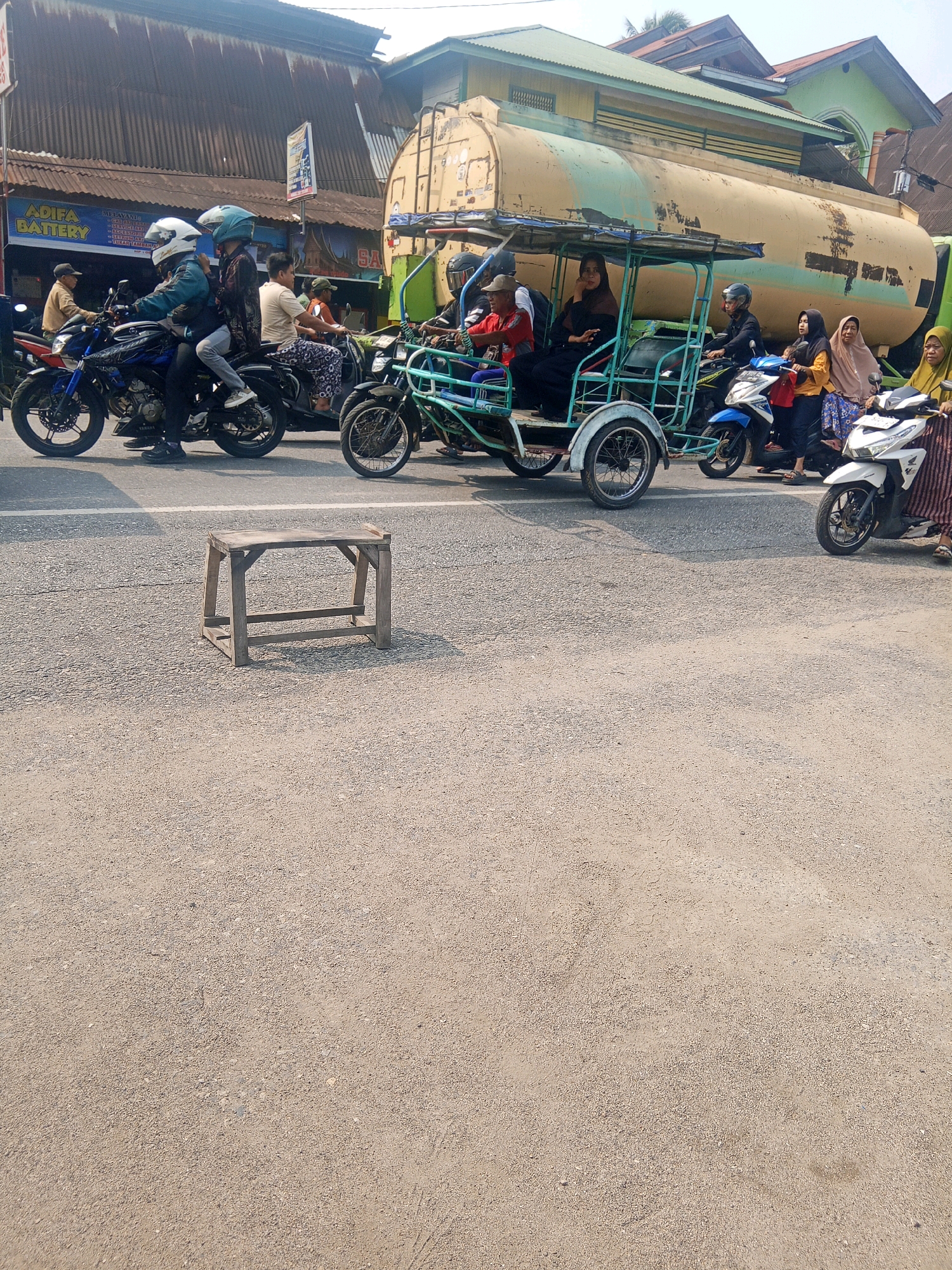 The image depicts a street scene in Nanggalo, Indonesia, characterized by a humid, sunny climate, with the sun casting distinct shadows on the asphalt road. In the foreground, a simple wooden stool sits abandoned on the road, its weathered surface suggesting it has been exposed to the elements.
The middle ground is a dynamic tableau of traffic, dominated by a fleet of motorcycles and a distinctive three-wheeled vehicle, possibly a becak motor. Several individuals are seen riding these vehicles, some wearing helmets, others not. The motorcycles are a mix of colors, with a prominent blue and black model in the left-center of the frame, and a white scooter on the right. A large, weathered tank truck, its yellow exterior faded and marked, occupies a significant portion of the background, its presence suggesting it might be a local delivery vehicle or part of the industrial landscape of Nanggalo.
The buildings lining the street are a blend of corrugated metal roofs and what appear to be more permanent structures, their colors muted by the bright sunlight. Signs are visible on the buildings, including one that reads "ADIFA BATTERY." People are dressed in light clothing, appropriate for the warm climate, with several women wearing hijabs. The overall impression is of a busy, everyday street scene in a tropical urban environment.