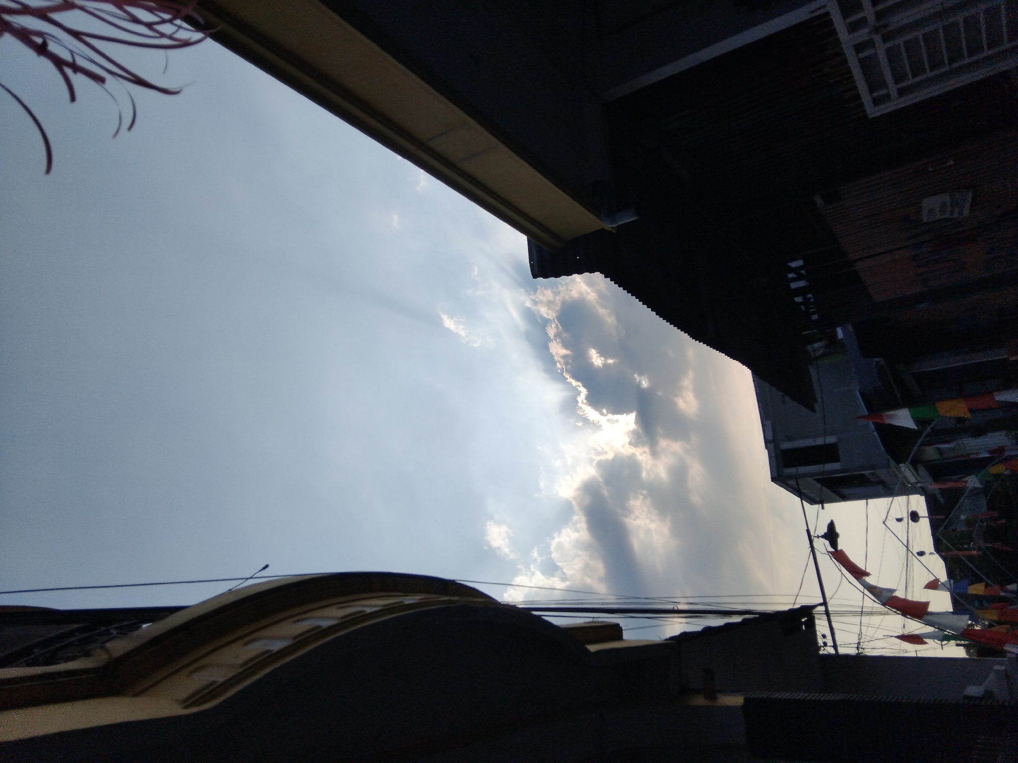 This is a low-angle, full-shot photograph of a residential street in Kepuh Kiriman, Indonesia, taken from a ground-level perspective looking upwards. The scene is dominated by the sky, which features a mix of bright sunlight breaking through clouds, creating visible sun rays, and a clear blue sky.
In the foreground and to the left, a dark, curved architectural element, possibly the side of a building or balcony with yellow trim, occupies a significant portion of the frame. To the right, another building's dark roofline with yellow trim angles upwards. Silhouetted against the sky are various residential structures, including a building with a corrugated metal roof and what appears to be a fence or gate on the far right.
Draped across the middle ground of the image are numerous small Indonesian flags and pennants in red and white, as well as multi-colored ones, strung across the street, indicating a possible celebration or event. A black lamp hangs from wires strung between buildings. The overall impression is of a slightly cluttered urban environment under a dynamic sky.