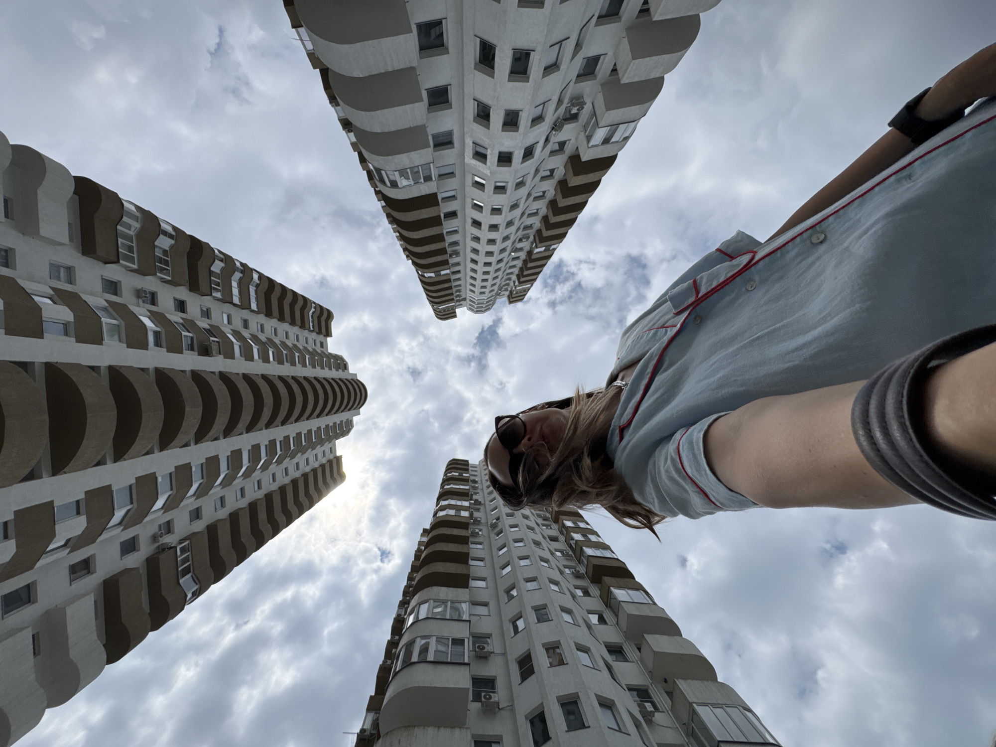 A low-angle, wide shot captures a woman taking a selfie with a fisheye lens in Sochi, Russia, looking up at the sky and surrounded by high-rise apartment buildings. The woman, with her hair blowing in the wind, wears sunglasses and a light blue pajama-style shirt with red trim. She holds a dark bag in her left hand. The architecture of the buildings, characterized by their curved balconies and white facades, creates a dramatic, converging perspective against a cloudy sky. The overall impression is one of urban grandeur and personal exploration within a city landscape.