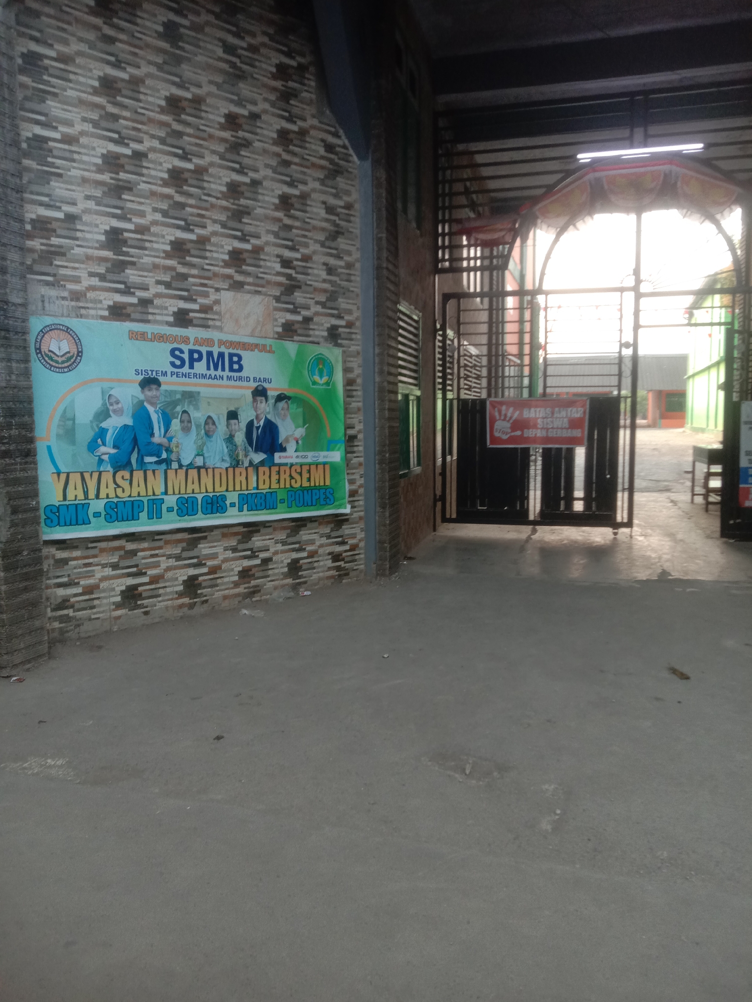 The image depicts the entrance of a school in Cianjur, Indonesia, as indicated by text on a banner. The banner, located on the left side of the image, advertises "SPMB SISTEM PENERIMAAN MURID BARU" and lists the affiliated institutions: "YAYASAN MANDIRI BERSEMI SMK - SMP IT - SD GIS - PKBM - PONPES". It features images of students and trophies.
To the right of the banner, a metal gate guards the school entrance. A red sign is attached to the gate, reading "BATAS ANTAR SISWA" and "STOP DEPAN GERBANG". Beyond the gate, the school grounds are visible, including buildings with colorful facades (green and red) and a metal canopy over an arched walkway. The ground in front of the gate is paved, and a shadow of the gate is cast on it. The overall environment appears to be daytime, with bright sunlight filtering through the gate. The wall to the left of the banner has a textured, tiled pattern.
