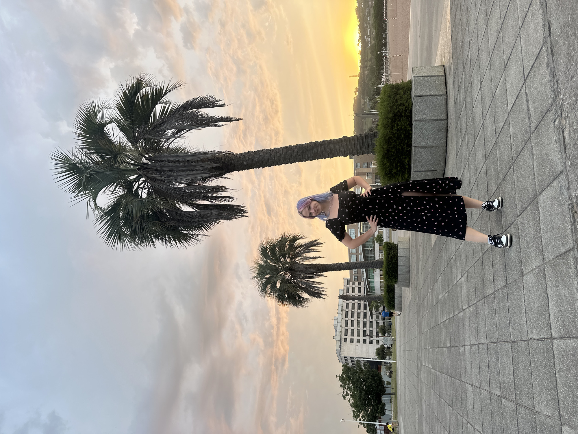 The image captures a young woman with lavender-colored hair standing on a paved plaza in Gijón, Spain. She is wearing a black, floral-patterned dress with a thigh-high slit and black and white sneakers. Her hands are placed on her hips, and she is looking at the camera. In the background, two palm trees stand tall, one larger and closer to the woman than the other. Beyond the trees, a building complex with white facades is visible, along with a beach and the ocean. The sky is filled with soft, pastel clouds, suggesting a sunset or sunrise. The overall setting appears to be a public space near the coast.