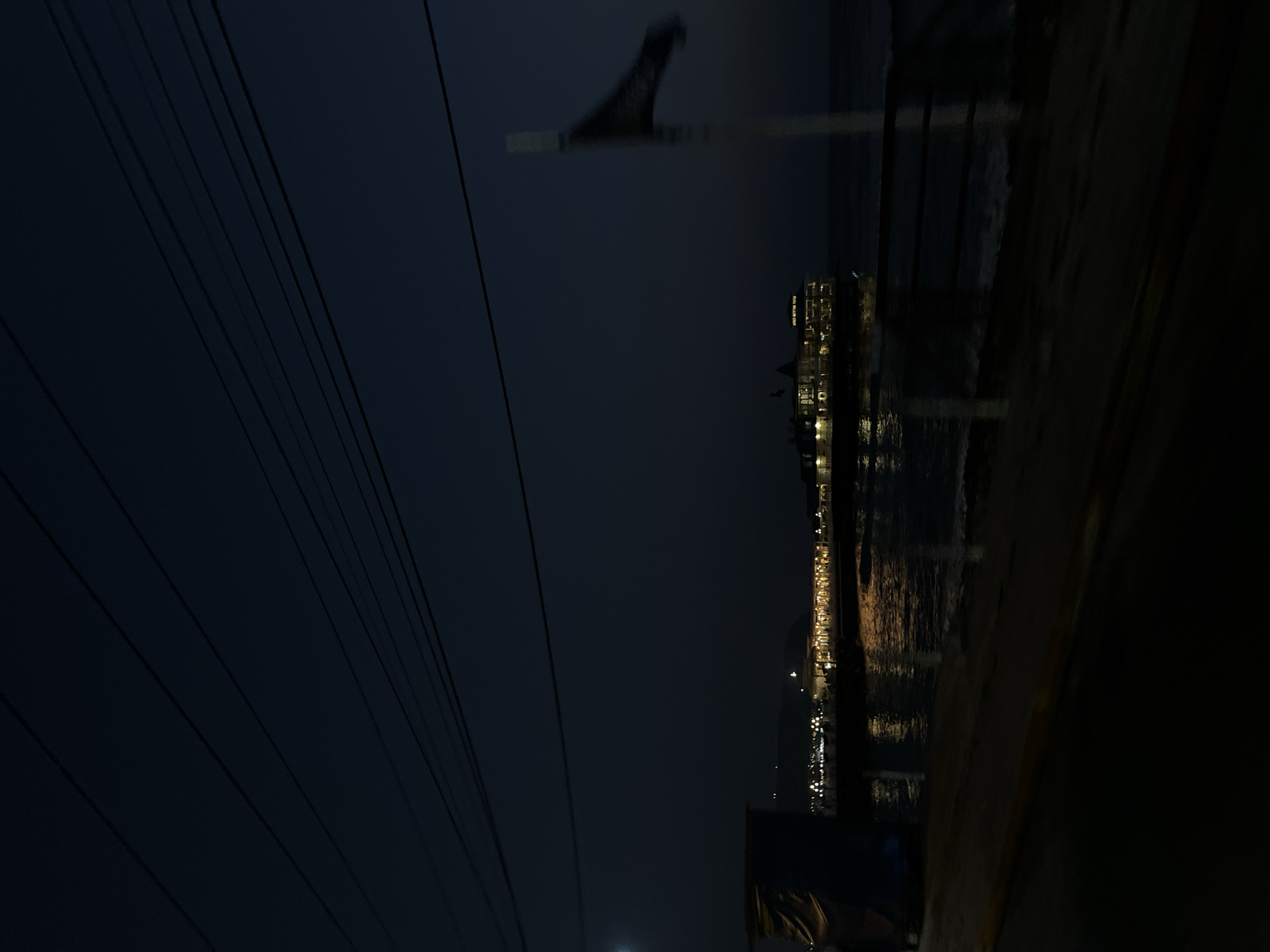 The image captures a nighttime scene in Miraflores, Peru, focusing on a brightly lit structure on a pier extending over the ocean. The sky is a deep, dark blue, with several parallel power lines stretching across the upper portion of the frame. The structure on the pier appears to be a building or restaurant, illuminated with warm, yellow lights, casting reflections on the dark, rippling water below. Small waves are visible along the shoreline in the foreground, which is dark and appears to be a sandy beach or promenade. A white pole with a dark, blurred flag is present on the right side of the image, partially obscuring the view of the pier and the ocean. A dark, nondescript structure, possibly a covered area or building, is visible on the far left. No people are clearly discernible in the image. The overall atmosphere is serene and nocturnal.