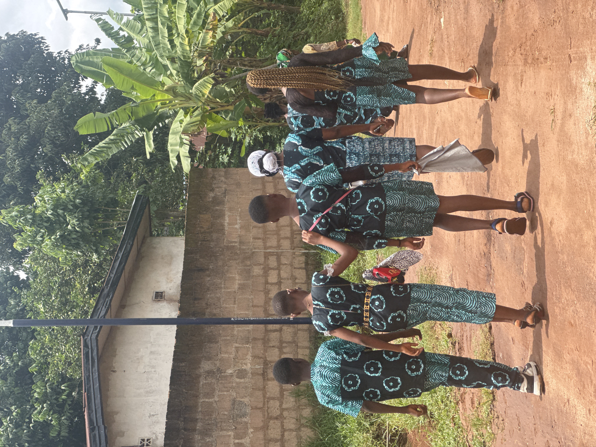 A group of five young women wearing matching dresses with a black background and light blue floral/circular patterns walk in a line in Eziudo, Nigeria. The dresses are knee-length with short sleeves. One woman has her hair in long braids. The environment shows a building with a brick wall, grassy areas, a banana tree, and other foliage. The ground is dirt and appears to be a path or road. There is an electricity pole in the background.