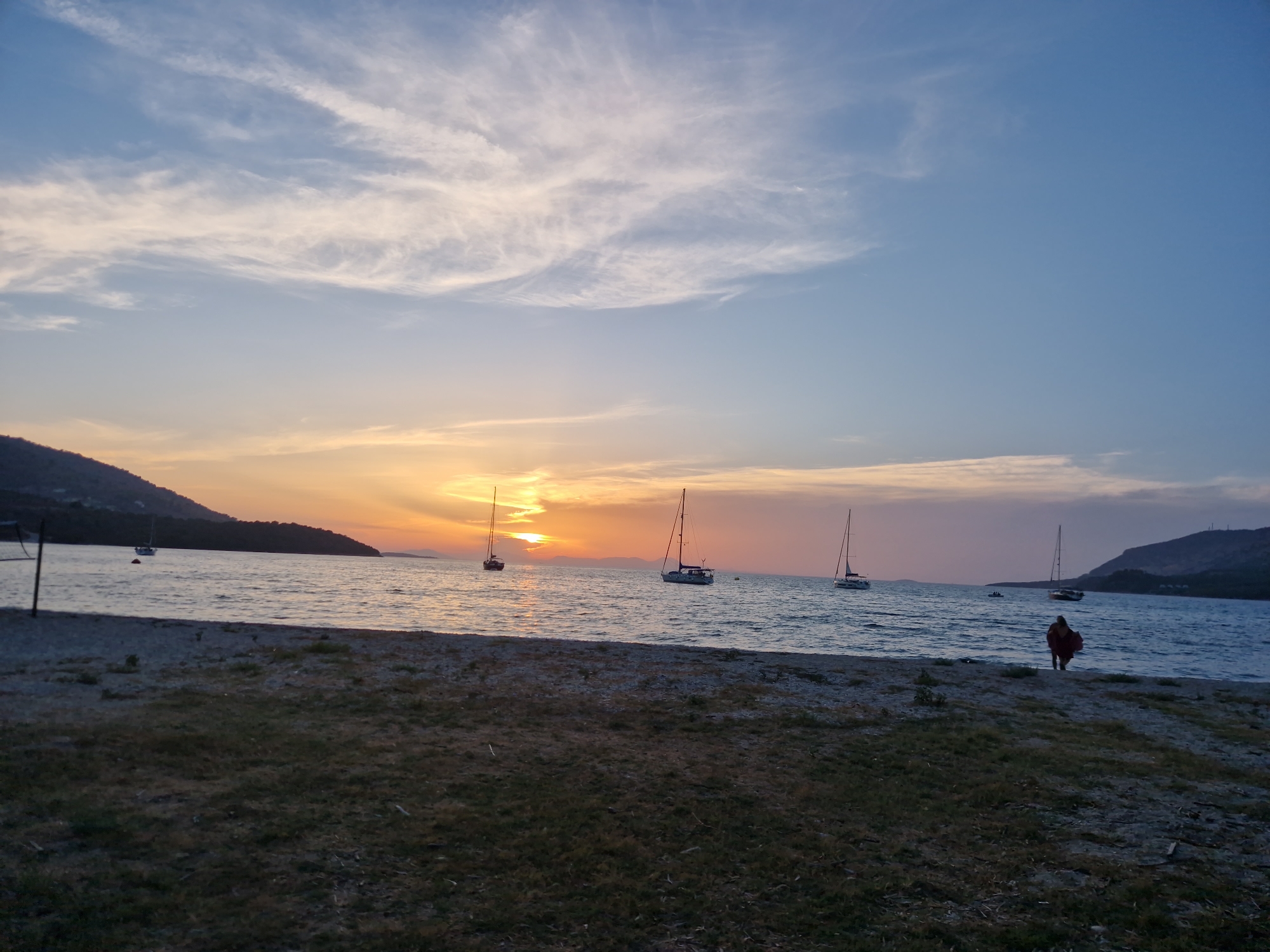 An outdoor, medium shot captures a sunset scene on a beach in Plataria, Greece. The sky is painted with hues of orange, yellow, and blue, with wispy clouds stretching across. In the distance, the sun is setting behind a mountain range, casting a warm glow on the water. Several sailboats are anchored in the calm sea. The foreground features a grassy, pebbly beach. A lone figure, wearing a red garment, is walking towards the water on the right side of the frame. The atmosphere is serene and peaceful.