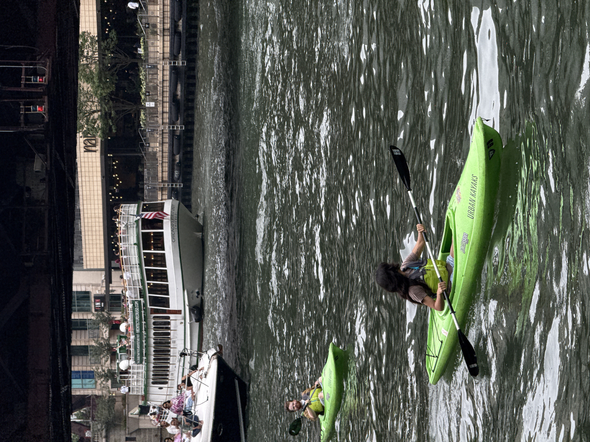 Two people are kayaking in the Chicago River, with a large tour boat and a building with "mai" signage in the background. The kayakers are in green kayaks, one with an "Urban Kayaks" logo. The tour boat, "Chicago's First Lady," is white with a green trim and has people on its upper deck. The building in the background has a beige facade and large windows. The river is dark and choppy, reflecting the overcast sky.