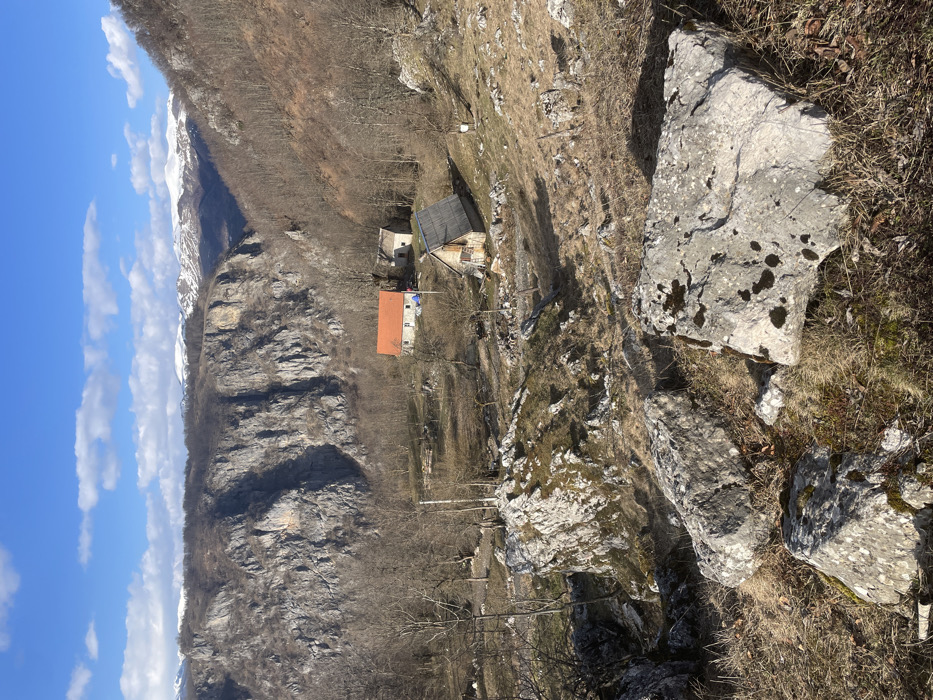 The image displays a mountainous rural landscape in Mokro, Montenegro, under a clear blue sky with scattered white clouds. The foreground features a dry, rocky incline populated by numerous large, light-grey boulders, some with moss patches, interspersed with sparse brown vegetation and dry grass. In the midground, a cluster of three structures is visible on a less steep slope. The primary structure is a single-story building with light-colored walls and an orange-tiled gable roof; a blue tarp is present adjacent to its base. Beside it, to the right, is a smaller, weathered structure with a dark, possibly wooden, gable roof. Below these, a third, smaller wooden building with a steeply pitched dark roof is situated. The midground also includes scattered bare deciduous trees. The background is characterized by steep, grey rocky cliffs and tree-covered mountainsides. Distant snow-capped mountain peaks are visible beneath the horizon. No individuals or active subjects are depicted. The scene is illuminated by natural sunlight, casting shadows.