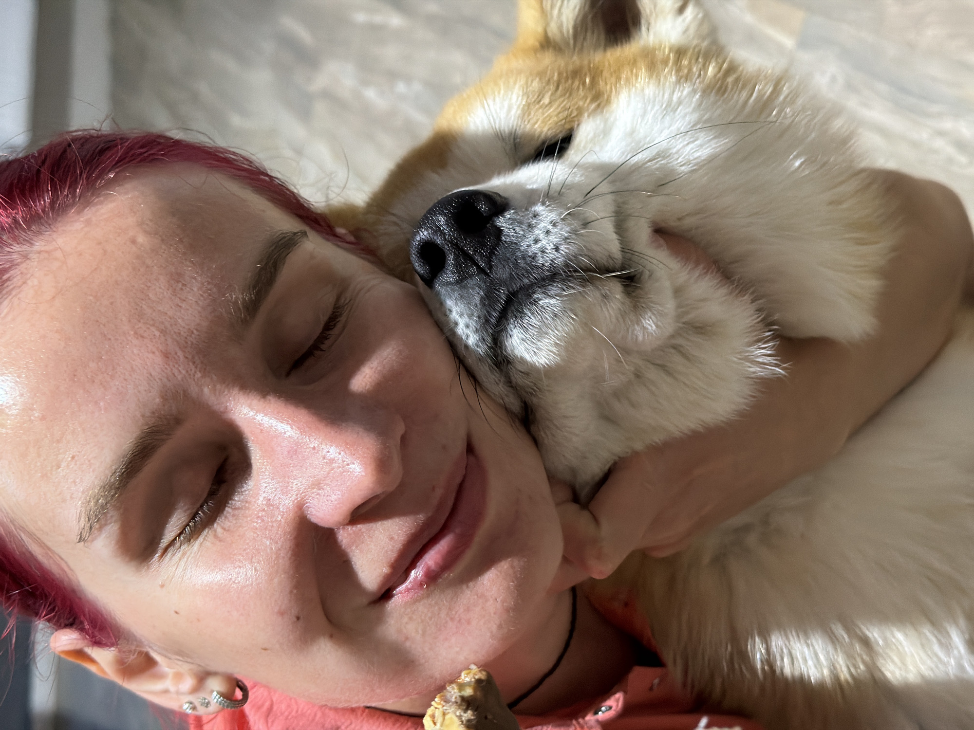 This is an indoor, close-up vertical shot depicting a person embracing a dog in the city of Chaikovsky, Russia. The person, appearing female, has dyed pink hair and fair skin with visible pores and minor blemishes. Her eyes are closed, and her lips form a gentle smile, suggesting contentment. She wears multiple silver earrings in her left ear and a light coral-colored top with a visible collar. A thin black cord is visible around her neck. Near the left side of her mouth, she holds a small, irregularly shaped brown object, possibly food. Her right arm is wrapped around a light-brown and white dog, likely a Shiba Inu, whose head is pressed against her cheek. The dog's black nose, closed eyes, and prominent whiskers are clearly visible. Both subjects appear calm and affectionate. The background consists of a light-colored, possibly tiled or laminated floor, illuminated by bright, directional light from the upper left.