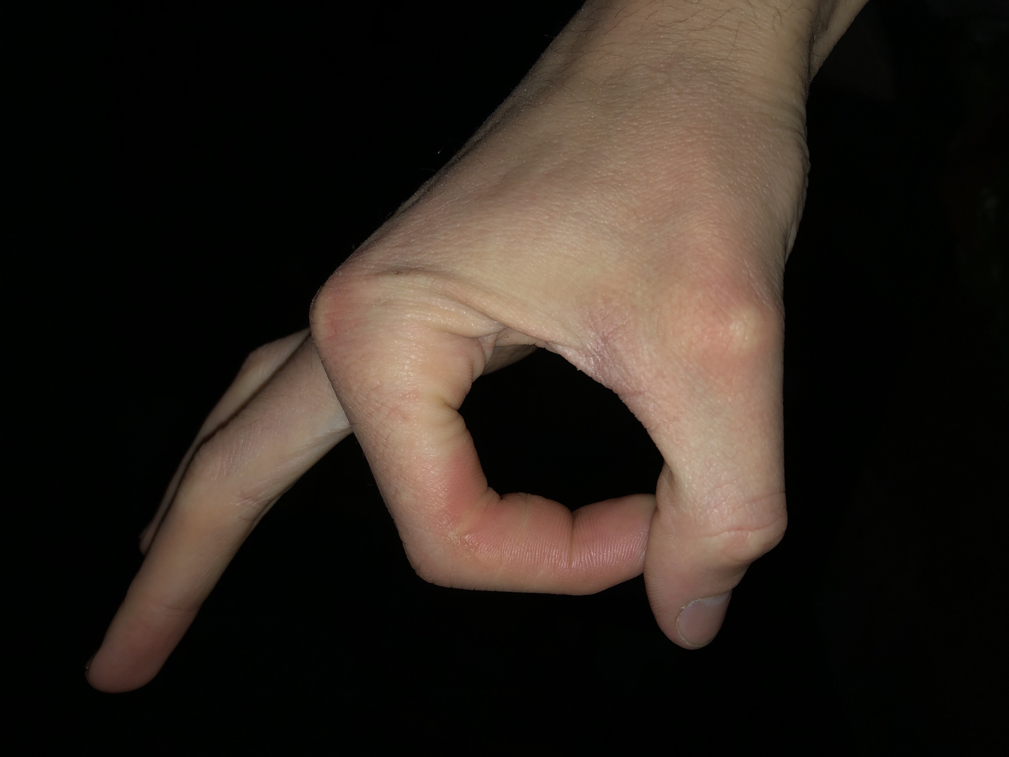 A Caucasian male hand is displayed against a dark background. The hand is making an "OK" gesture, with the thumb and index finger forming a circle and the remaining fingers extended. The location is unknown.