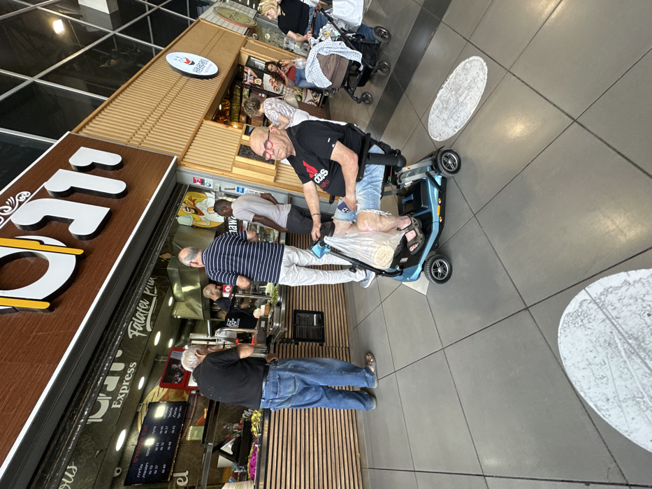 The scene depicts an active food court area within an indoor commercial complex in Mishmar Hasharon, Israel. The foreground features a bald, older male with red-rimmed glasses, wearing a black Adidas t-shirt, ripped light blue denim shorts, and dark sandals, seated in a blue and black four-wheeled electric mobility scooter. A clear plastic bag containing flatbreads hangs from the scooter's handlebars, and his right leg displays visible scarring or discoloration. He is looking directly forward. The floor is composed of large, light grey tiles, with two distinct oval-shaped, light-colored textured patches visible in the lower frame.
In the midground, a food stall identified by a large brown sign with Hebrew text and the English words "Express Falafel Kun" and "Shawarma" is prominently featured. The stall has a light brown slatted wooden counter behind which food items, including salads and sauces, are displayed. Two male employees are visible behind the counter, one in a black t-shirt is actively preparing food. Three male customers are gathered at the counter: an older man with grey hair in a black shirt and blue jeans stands to the left; an older man in a striped shirt and light trousers stands in the middle, facing away; and a younger man with dark skin in a light grey t-shirt stands to the right. All three customers are observing the food preparation or waiting to order.
The background reveals additional elements of the commercial space. To the right of the main food stall, another stall with a light wooden facade and a white circular "Hello Baby" logo is visible. Several individuals are present in the background, including two women standing near the second stall, one in a light top and another in a red top. Further right, a woman with blonde hair in a black top stands beside a brown baby stroller containing a blanket or clothing. The upper background shows a high ceiling with a modern, possibly glass-paneled architectural structure, indicative of a large indoor complex.