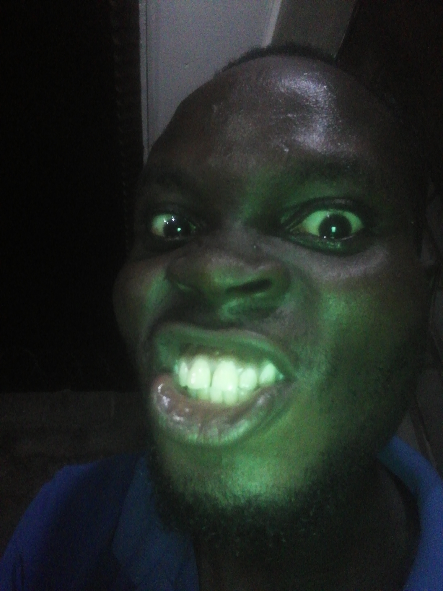 The image shows a close-up of a dark-skinned person's face with a green cast. The person is grimacing, with teeth bared. The background is dark and indistinct. There is no evidence in the image to suggest that this image was taken in Ikorodu, Nigeria.