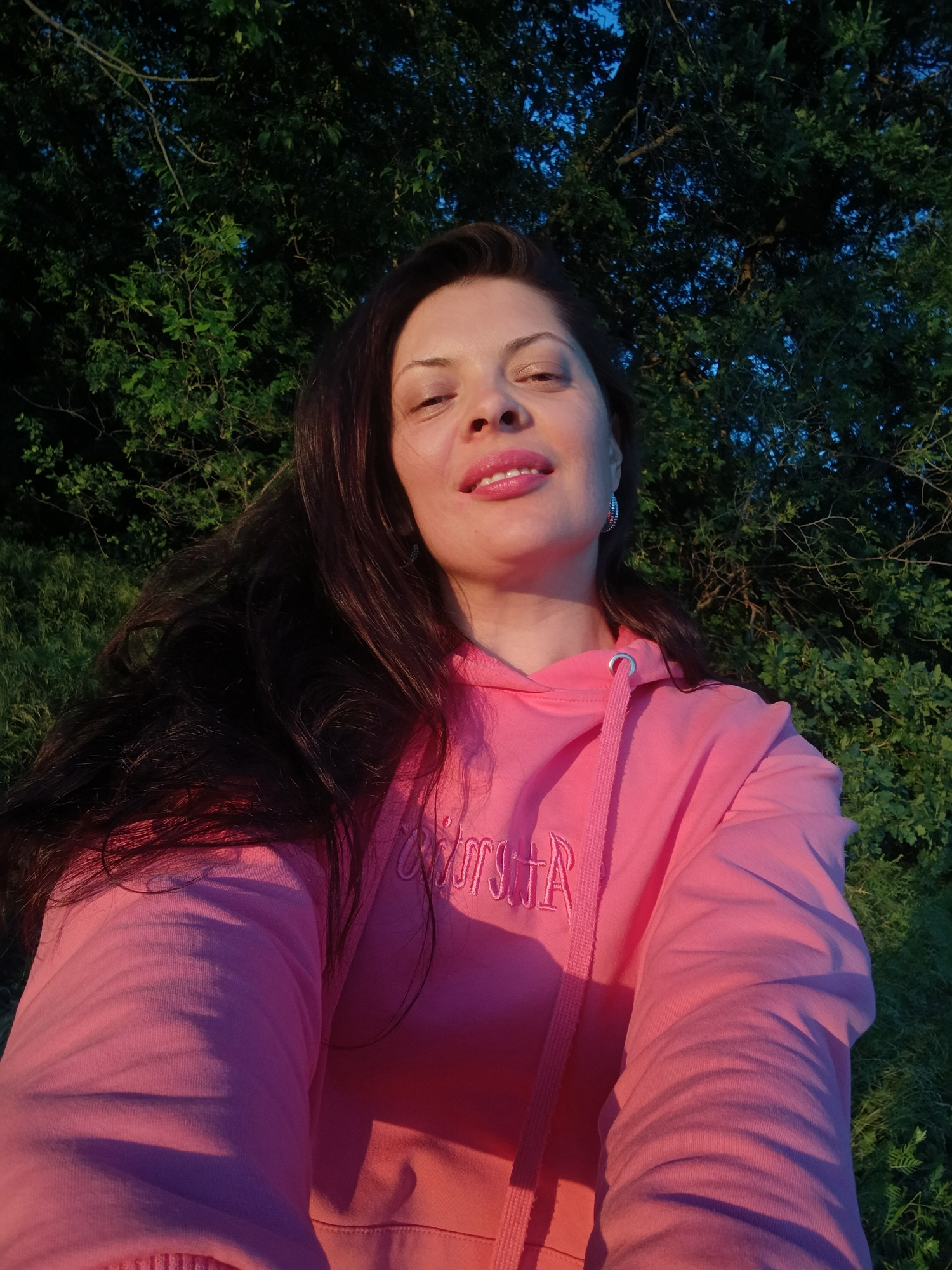 A woman with long, dark hair and pink lipstick is taking a selfie outdoors. She is wearing a pink hooded sweatshirt with the word "ROXY" embroidered on the chest in white. The background consists of green foliage and a dark blue sky, suggesting an evening setting. The lighting indicates the sun is low in the sky, casting a warm glow on the subject. The scene is likely a park or natural area within the city of Saratov, Russia, given the presence of trees and grass.