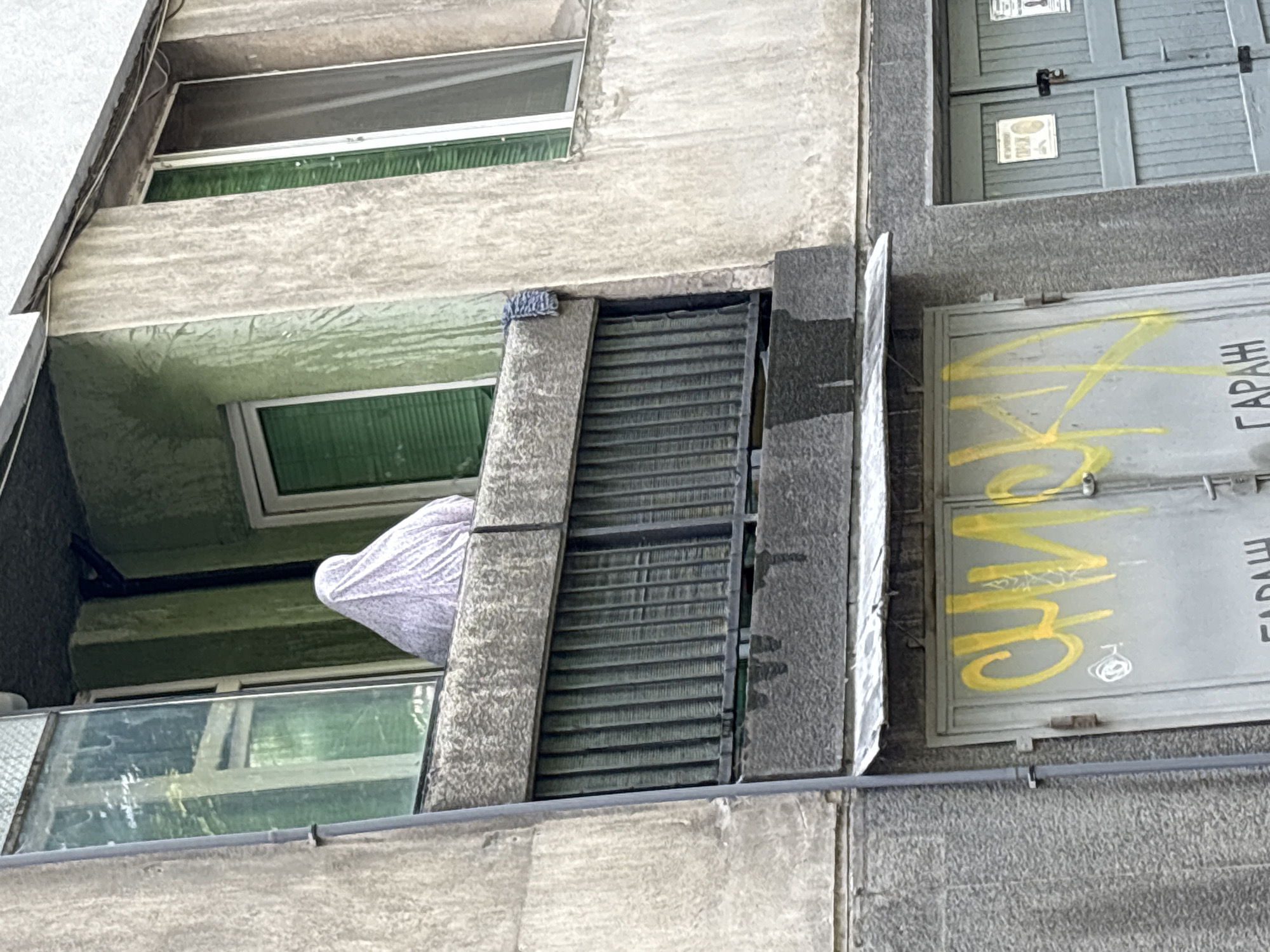 A medium shot, taken at an oblique angle, depicts the exterior of an urban building in Plovdiv, Bulgaria. The building appears to be constructed of heavily textured, light-grey stone or concrete. Several windows are visible, some obscured by either grilles or reflective glass. A white, somewhat amorphous object, possibly fabric, is partially visible hanging over the edge of an upper-story balcony, situated between two windows. The corner of a nearby garage door, bearing yellow graffiti, is also visible.
