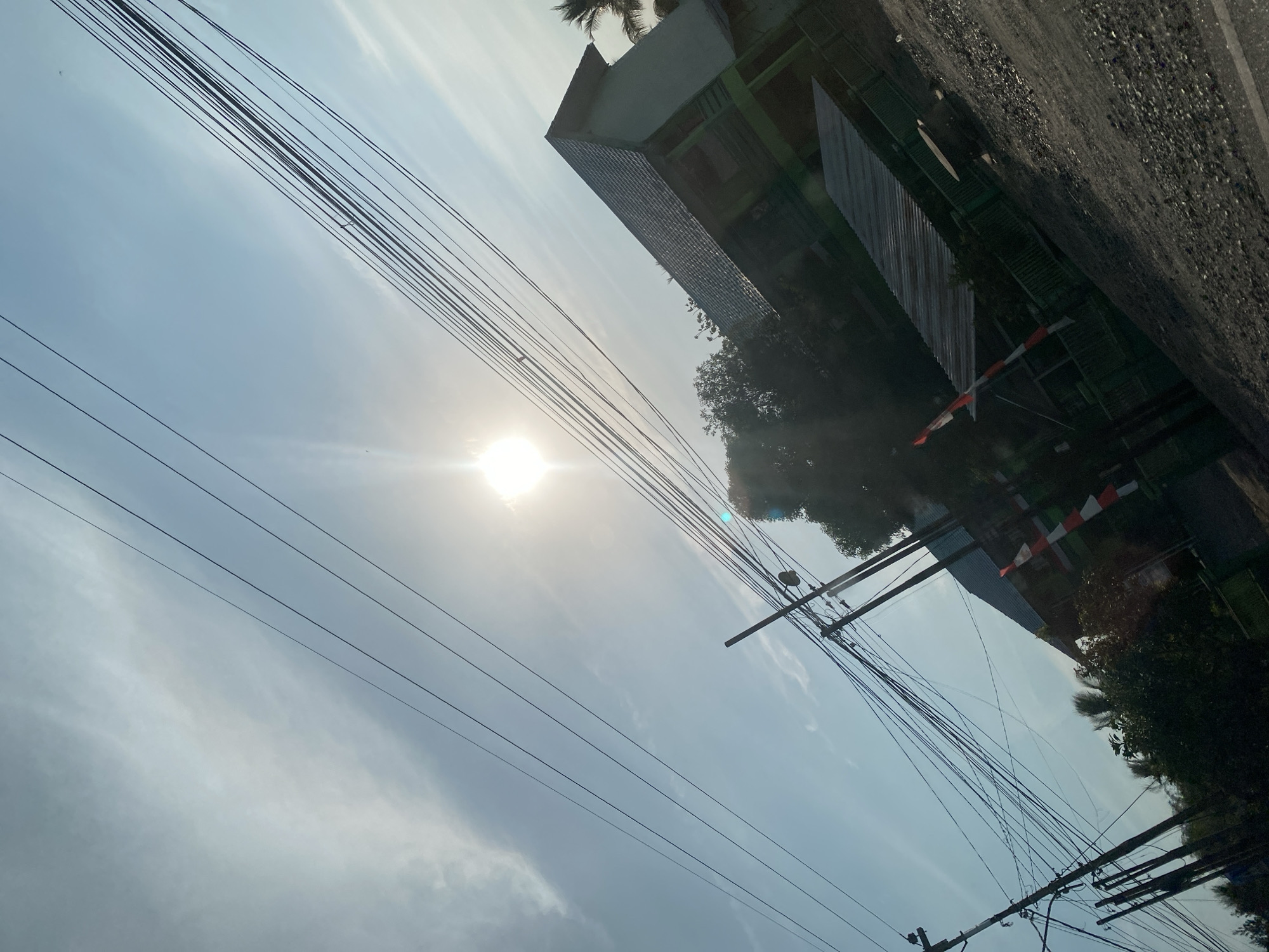 The image depicts a street scene in Astambul Kota, Indonesia, under a bright, hazy sky with the sun prominently featured. Power lines stretch diagonally across the frame, creating a sense of depth and visual complexity. Several utility poles with numerous wires are visible. In the background, a building with a gray, tiled roof and green accents is partially obscured by trees. In front of this building, a red and white striped barrier or pole is present, possibly indicating a checkpoint or a marker. The ground appears to be paved with gravel and asphalt, with some vegetation bordering the area. The overall impression is of an outdoor environment, possibly a residential or semi-urban area, during daylight hours.