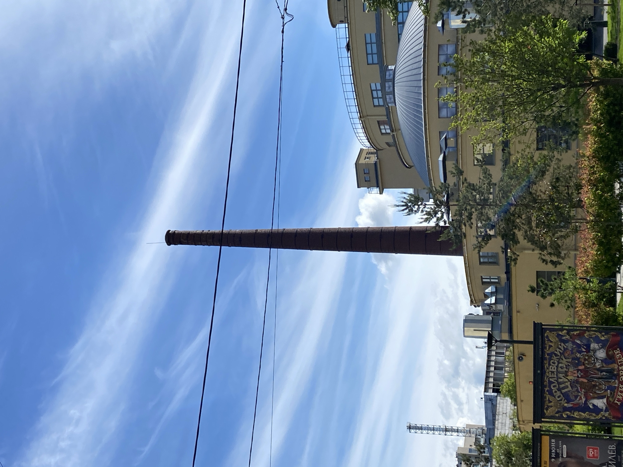 A low-angle photograph shows a tall brick chimney extending vertically against a blue sky with cirrus clouds in Saint Petersburg, Russia. Power lines run in parallel to the chimney. To the right of the chimney is a building with a rounded roof, trees, and a street-level sign with Cyrillic text. In the background, a crane and other buildings are visible.