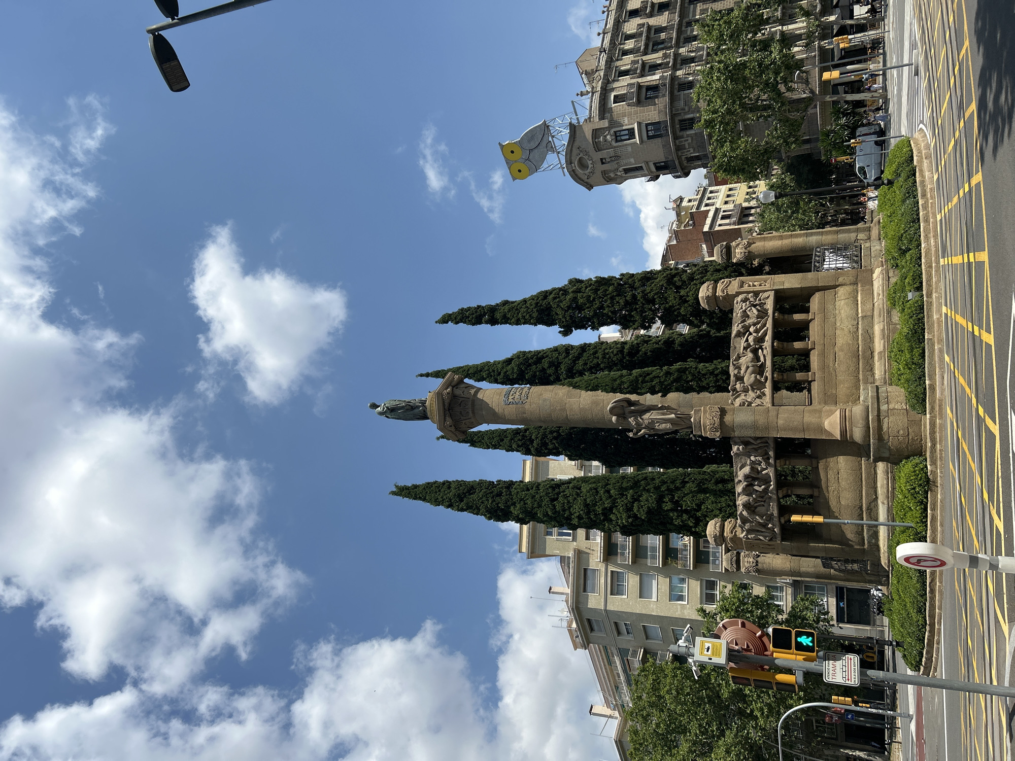 This image shows the Monument a Colom (Columbus Monument) in Barcelona, Spain, from a street-level perspective. The monument is surrounded by tall, thin evergreen trees. It is a stone structure with carvings. A statue stands atop the column. The sky is blue with scattered white clouds. In the background are buildings of varying heights, with the "Casa de la Lactancia" building topped with an Owl (El búho) advertisement visible in the upper part of the image. A street and traffic lights are in the foreground, showing yellow striped markings, and a pedestrian crossing with a green light illuminated.