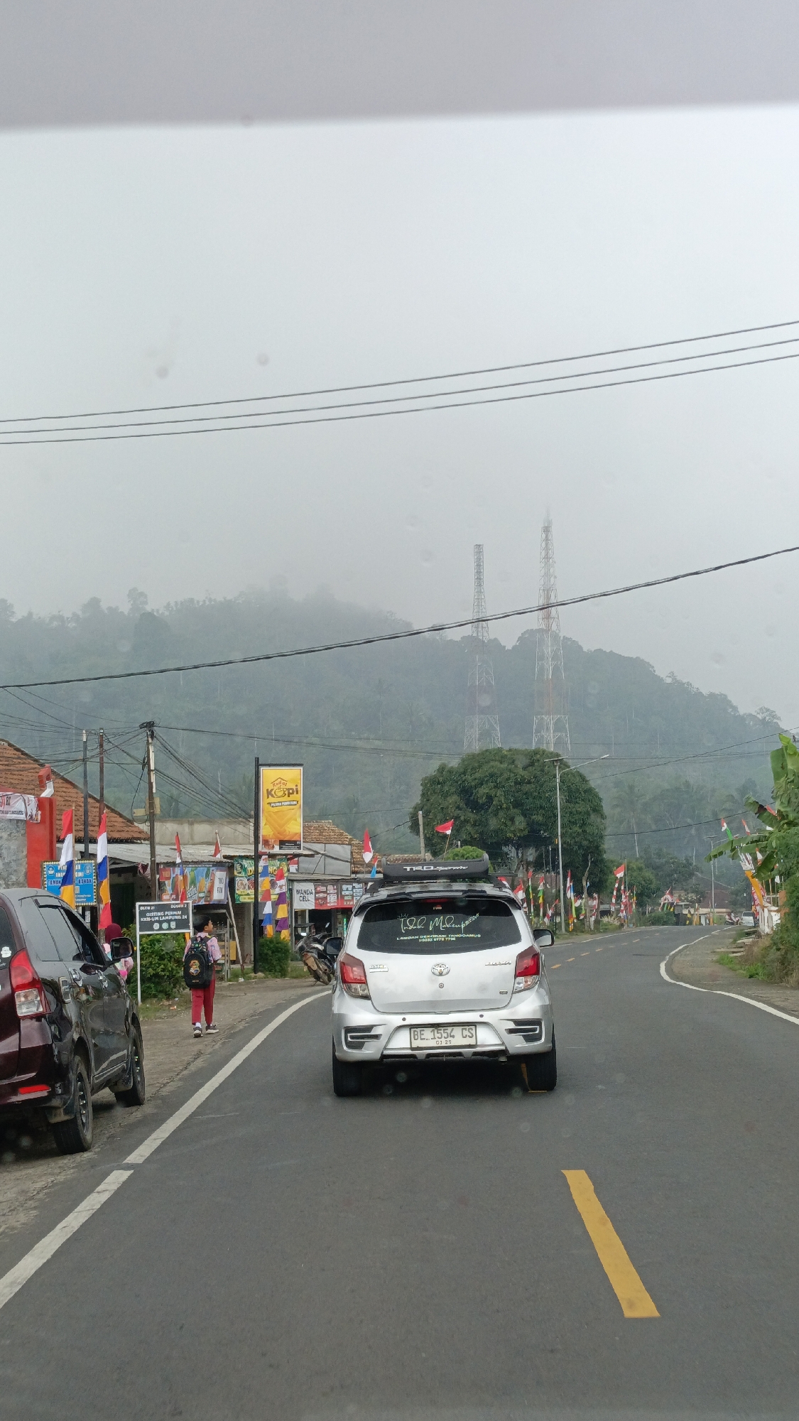 The image depicts a road scene in Sukabanjar, Indonesia, on a hazy day. In the foreground, a silver car with license plate BE 1554 CS is driving on the right side of the road. A dark red car is parked on the left side of the road. A person wearing a pink shirt and red pants with a backpack is walking on the left side of the road. Businesses with flags and signs are visible along the roadside, including a yellow sign with the word "KOPI". In the background, two tall radio towers stand against a backdrop of misty green hills covered in trees. The overall atmosphere is overcast, with a sense of a rural or semi-urban environment.