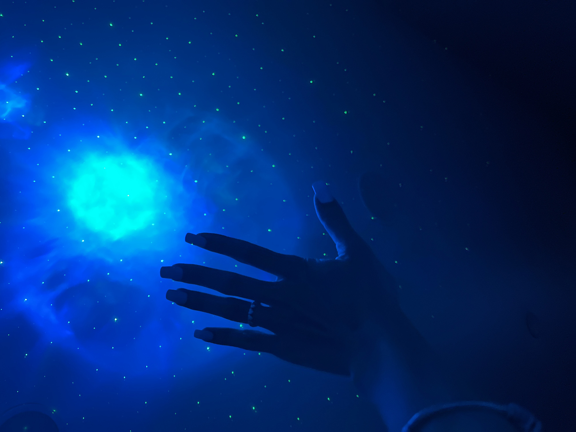 The image shows a person's hand reaching out towards a projection of a nebula and stars, which illuminates a dark blue room. The hand is silhouetted and has a ring on one finger. The projection is a bright, swirling blue and green nebula with scattered green dots representing stars. The lighting creates a mystical or celestial atmosphere. The location is not discernible from the image; there is no information provided about Nizhny Novgorod, Russia.