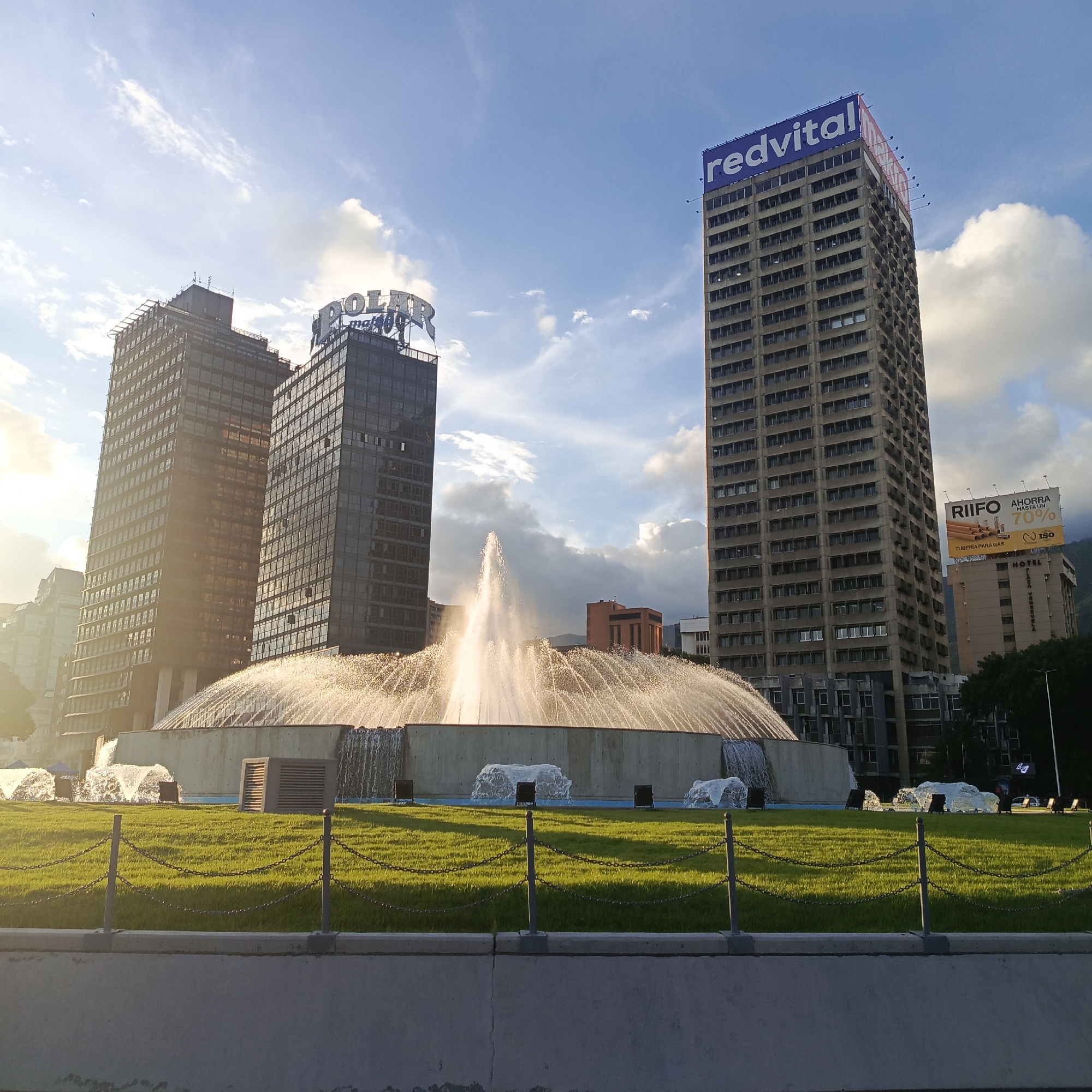This is a medium shot, eye-level, horizontal photograph of the Plaza Venezuela fountain in Caracas, Venezuela. The fountain is the central focus, with water shooting high into the air, illuminated by the setting sun, which casts a warm, golden light on the scene. The sun also highlights the lush green grass in the foreground, separated from the fountain by a low concrete barrier and chain-link fence.
Behind the fountain, three tall modern buildings dominate the background. The building on the far left is dark and reflective. The middle building is also dark and features a large, illuminated sign that reads "POLAR." The building on the far right is a lighter concrete color and has a prominent blue sign at the top that says "redvital." Further to the right, a lighter-colored hotel building is visible, with a billboard above it advertising "RIIFO AHORRA HASTA UN 70% TUBERÍA PARA GAS."
The sky is a vibrant blue with scattered white clouds, suggesting fair weather. The lighting indicates it is late afternoon or early evening, likely during a period of golden hour. The overall mood is serene and majestic, showcasing an urban landmark with natural beauty. There are no people visible in the frame. The image is a still capture of the fountain and surrounding cityscape.