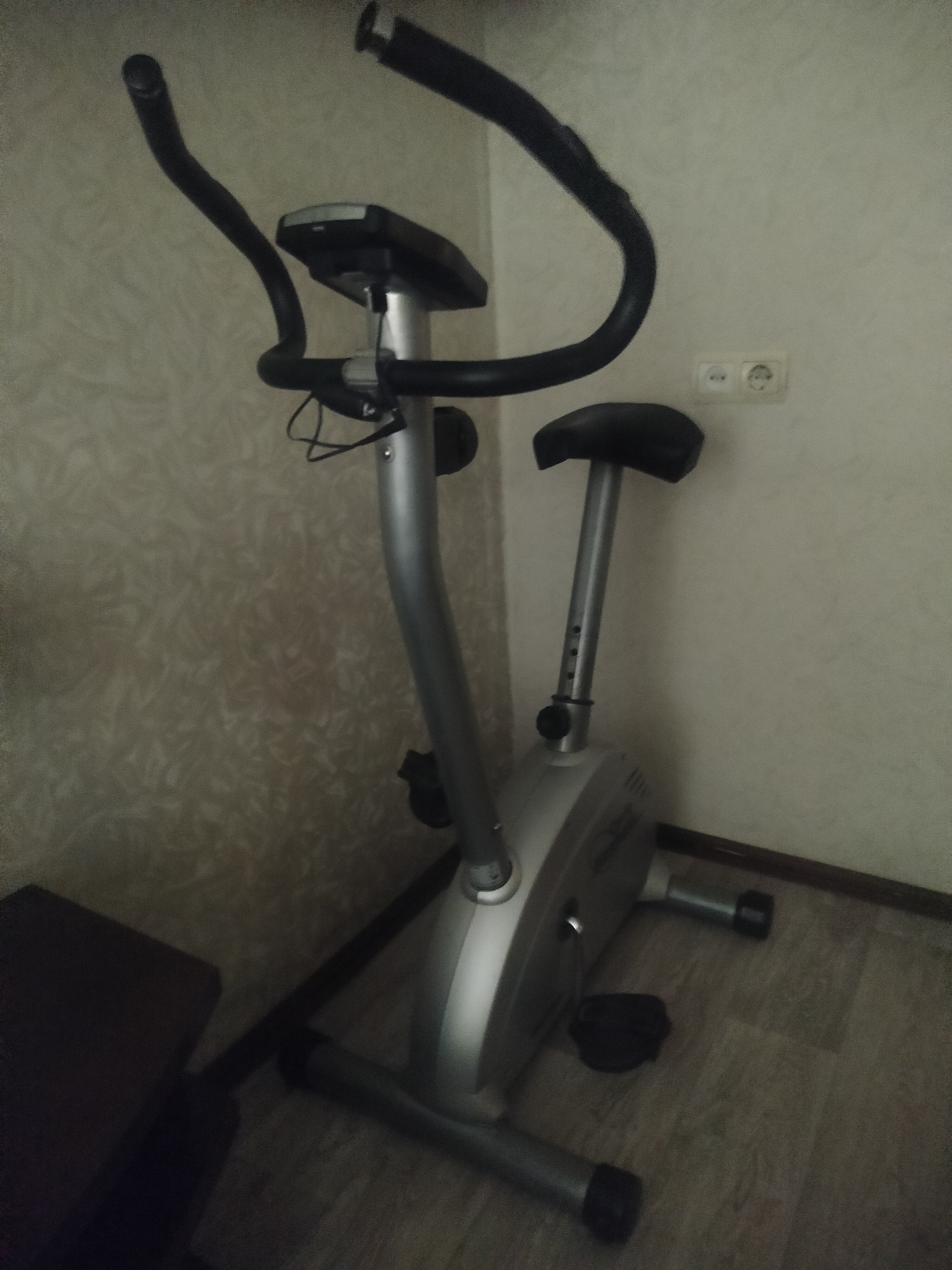 The image shows a stationary exercise bike indoors, positioned against a wall with a floral or textured wallpaper. The bike is primarily silver and black, featuring a seat, handlebars with integrated monitor, and pedals. An electrical outlet is visible on the wall behind the seat. The floor appears to be linoleum or vinyl. The presence of a dark object on the left side of the frame suggests the edge of another piece of furniture or equipment. There is no way to determine the specific location within the city of Irkutsk, Russia, or the presence of any people.