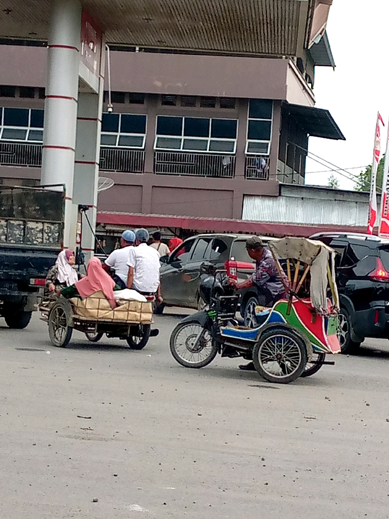The image depicts a street scene in Keudebayu, Indonesia, with a focus on two motorized tricycles, commonly known as "becak motor." In the foreground, a becak motor with a colorful red, green, and blue painted body is being operated by a man wearing a patterned shirt and a dark cap. The becak motor has a covered passenger area with yellow fabric. Beside it, another becak motor is carrying two women, one in a patterned headscarf and dark clothing, and another woman draped in a pink headscarf, sitting in the passenger compartment which appears to be constructed from woven material. They are seated on what seems to be goods. Behind these becak motors, several cars are visible, including a black SUV and a dark-colored sedan. In the background, a building with a corrugated metal roof and multiple windows is present. The street surface is paved and appears somewhat dusty. Flags are visible on the right side of the image, suggesting a public area or event. The overall impression is a snapshot of daily life and transportation in a street environment.